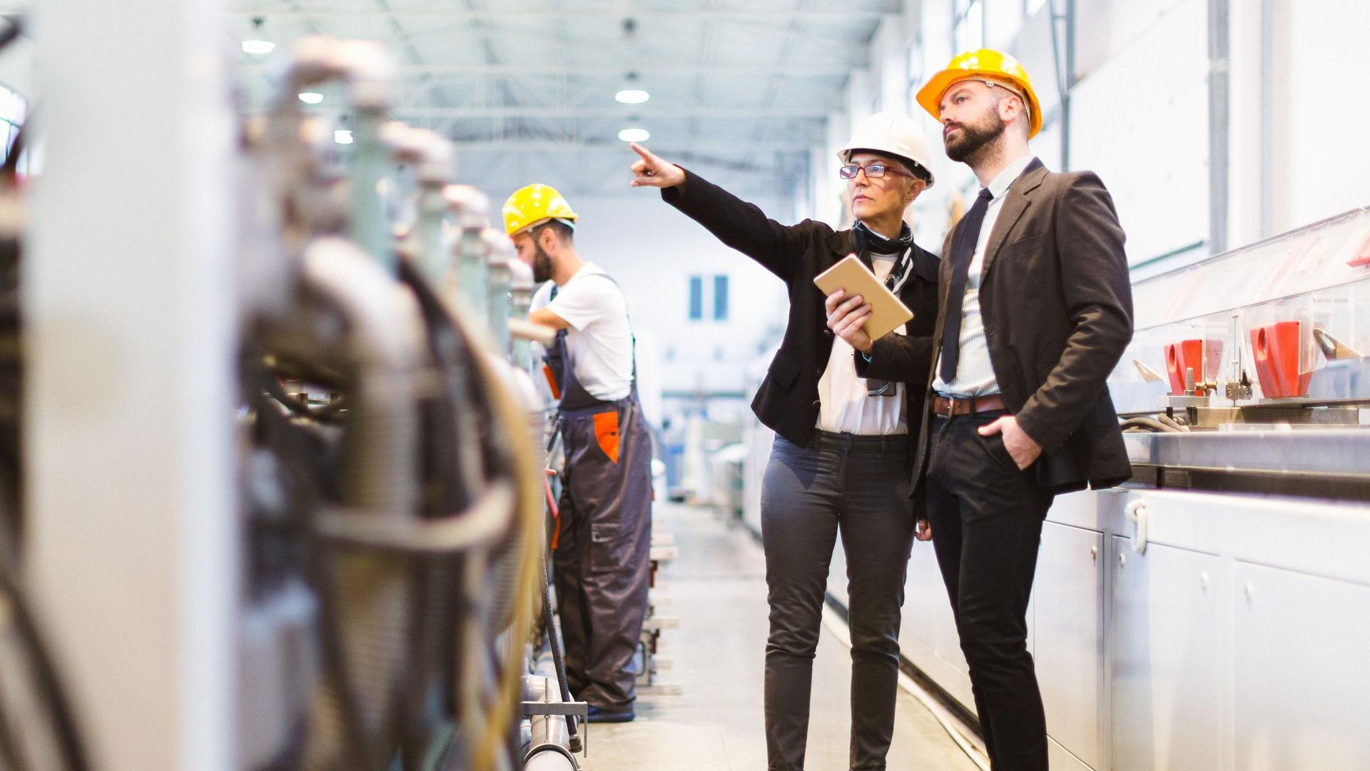 Eine Frau und ein Mann in Business-Kleidung stehen mit Helmen in einer Fabrik. Im Hintergrund arbeitet ein Mann an einer Maschine.