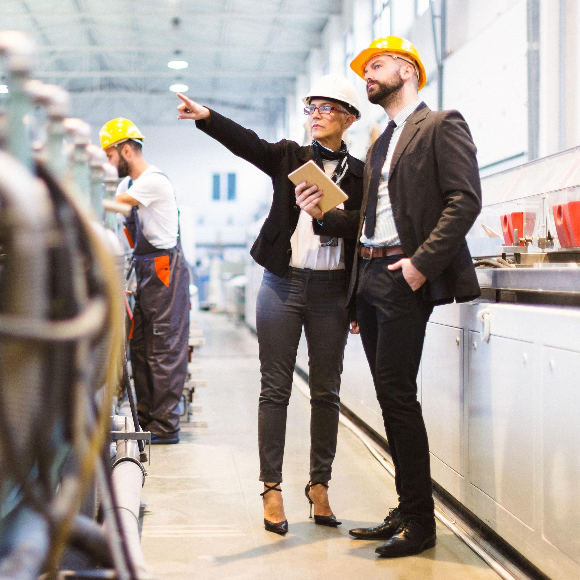 Eine Frau und ein Mann in Business-Kleidung stehen mit Helmen in einer Fabrik. Im Hintergrund arbeitet ein Mann an einer Maschine.