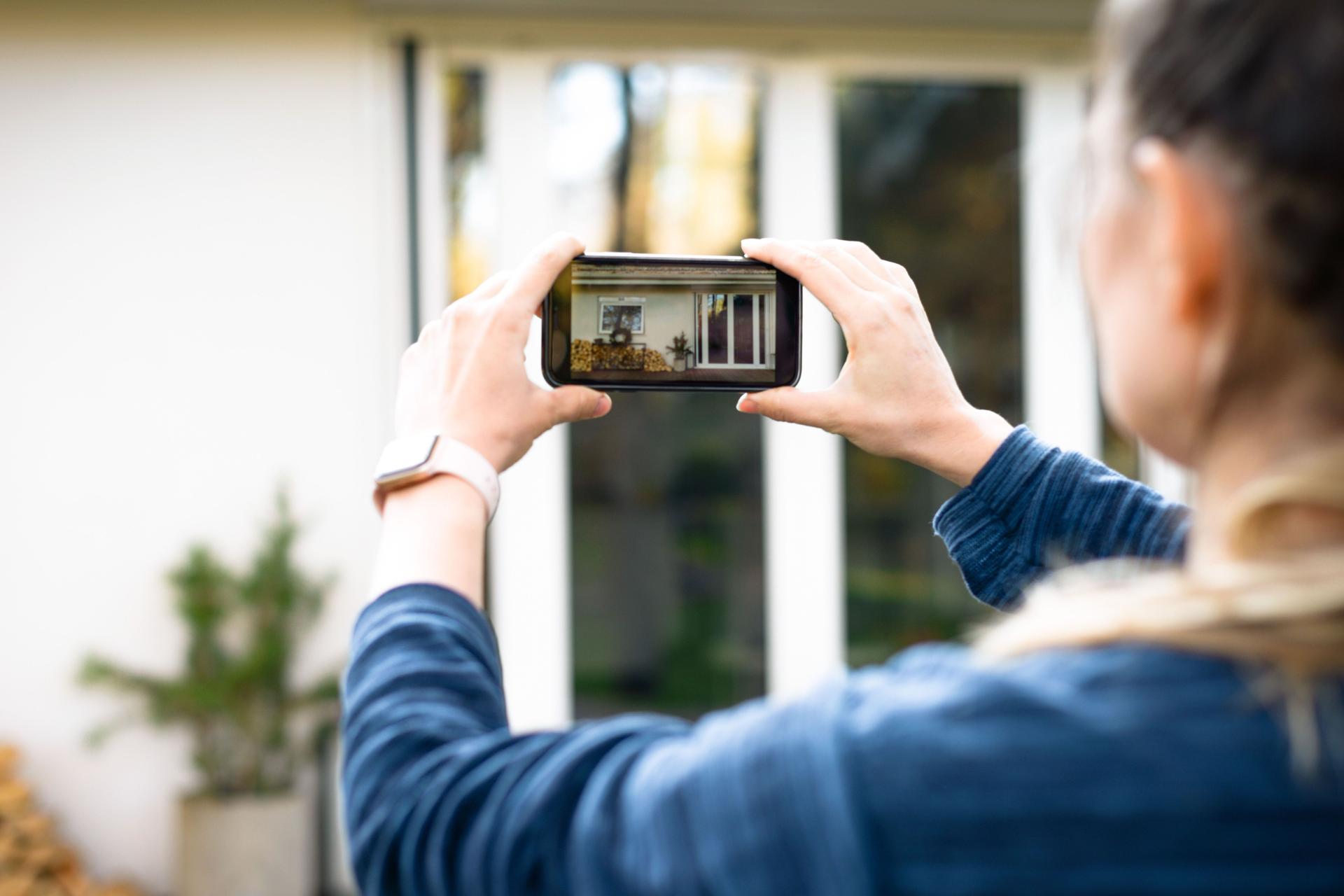 Eine Frau fotografiert mit ihrem Smartphone die Terrassentür eines Hauses von außen. Das Foto wird dabei auf dem Smartphone-Bildschirm angezeigt.