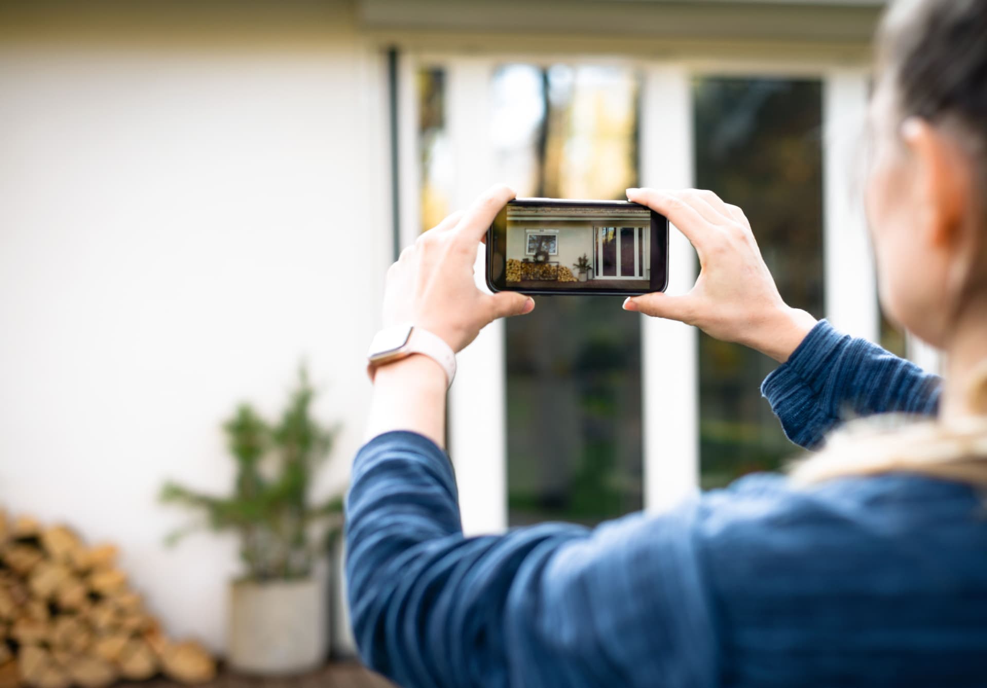 Eine Frau fotografiert mit ihrem Smartphone die Terrassentür eines Hauses von außen. Das Foto wird dabei auf dem Smartphone-Bildschirm angezeigt.