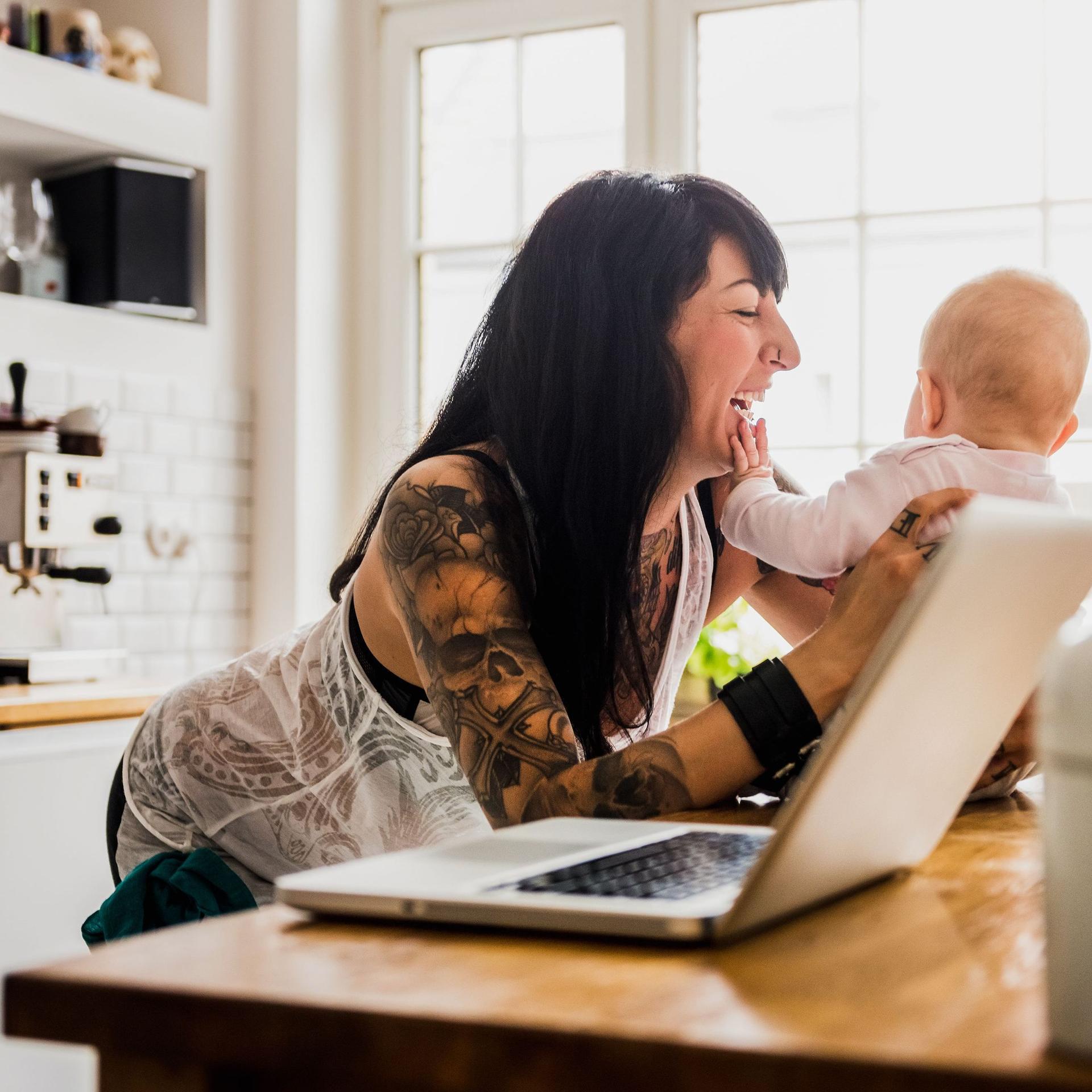 Eine junge tätowierte Mutter mit Baby in einer modernen hellen Küche. Neben ihr ist ein Laptop. Das Baby greift in das lachende Gesicht der Mutter.