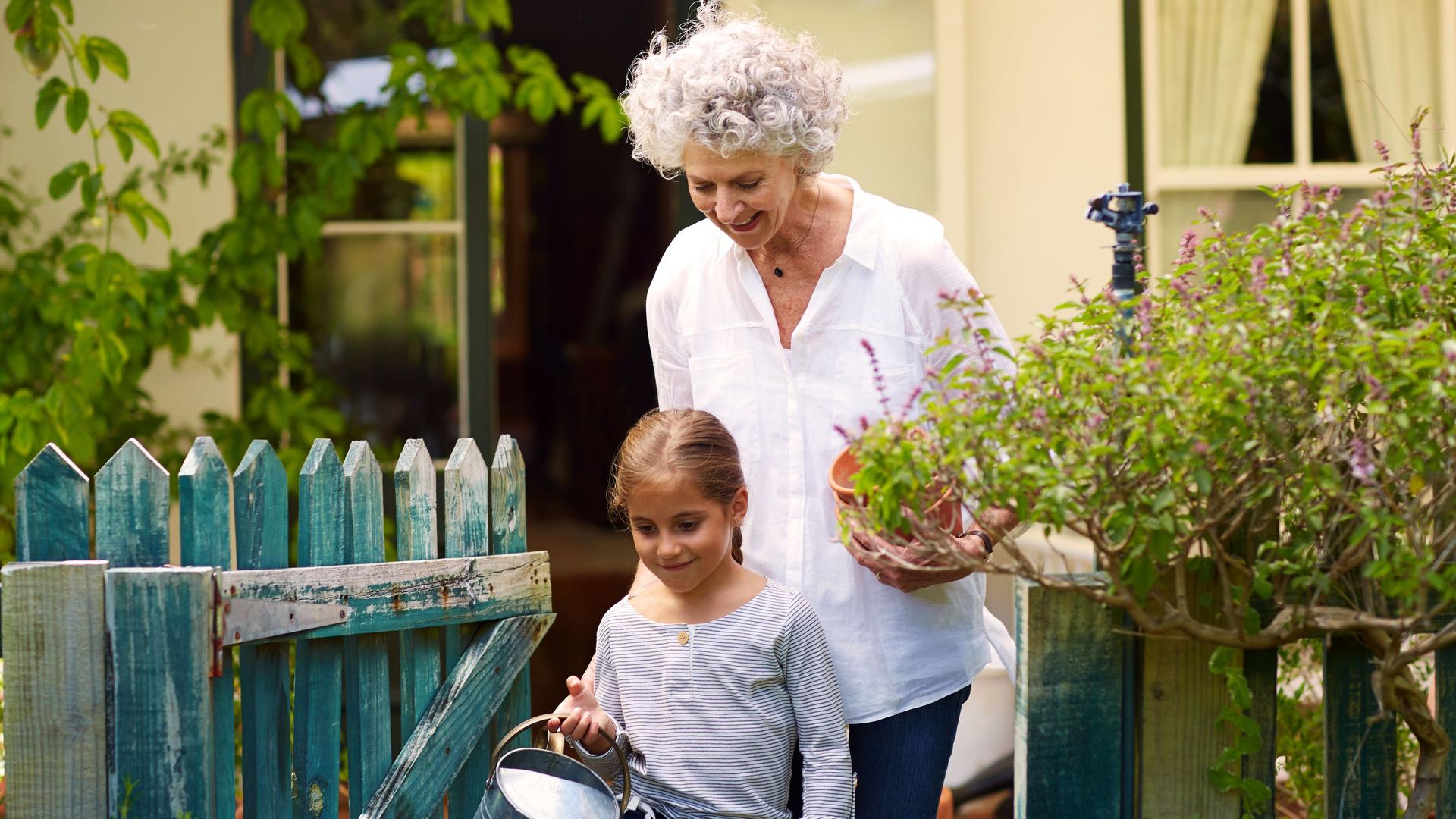Oma und Enkelin haben Gartenarbeitsutensilien in den Händen und laufen durch ein Gartentor. Im Hintergrund ist ein Einfamilienhaus zu sehen.