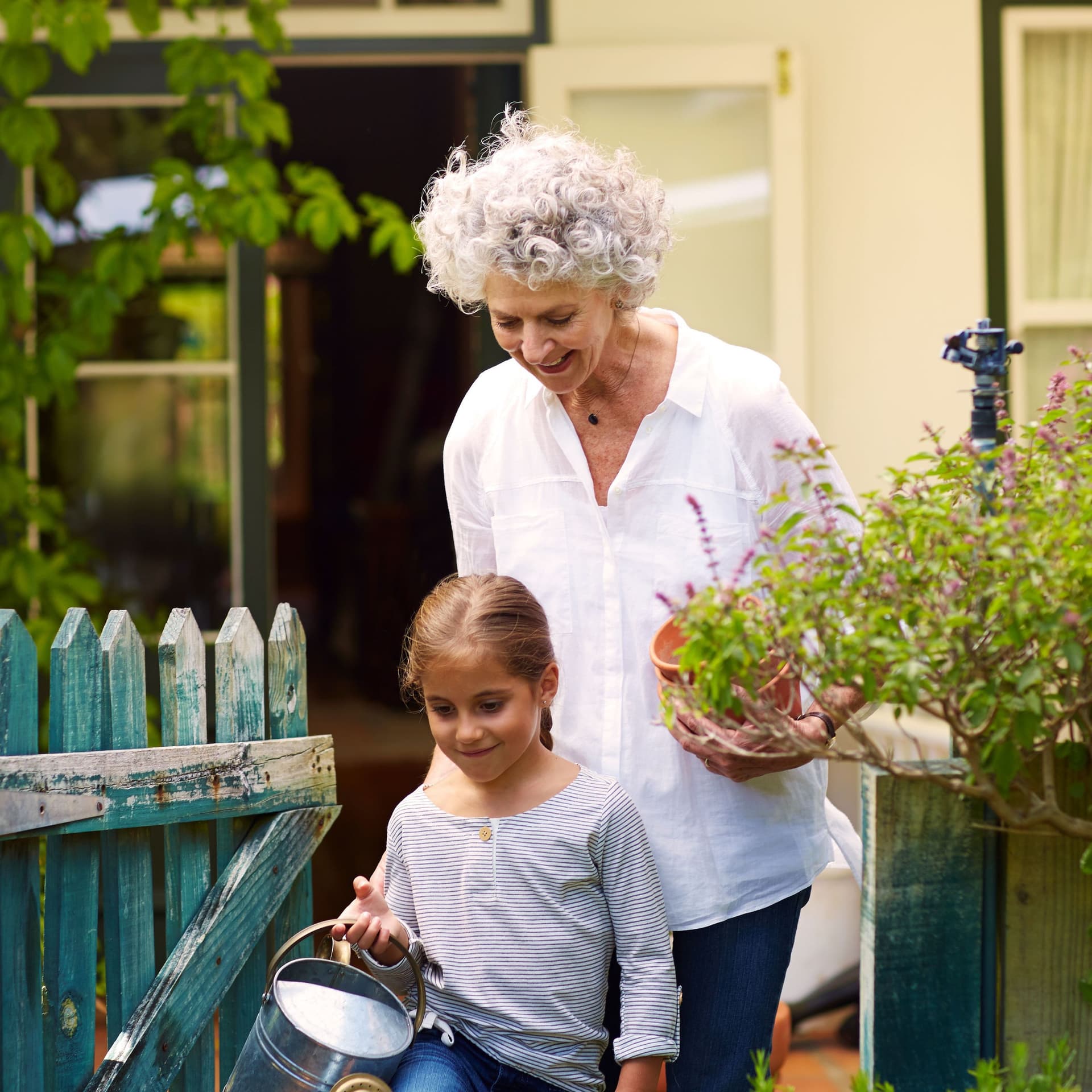 Oma und Enkelin haben Gartenarbeitsutensilien in den Händen und laufen durch ein Gartentor. Im Hintergrund ist ein Einfamilienhaus zu sehen.