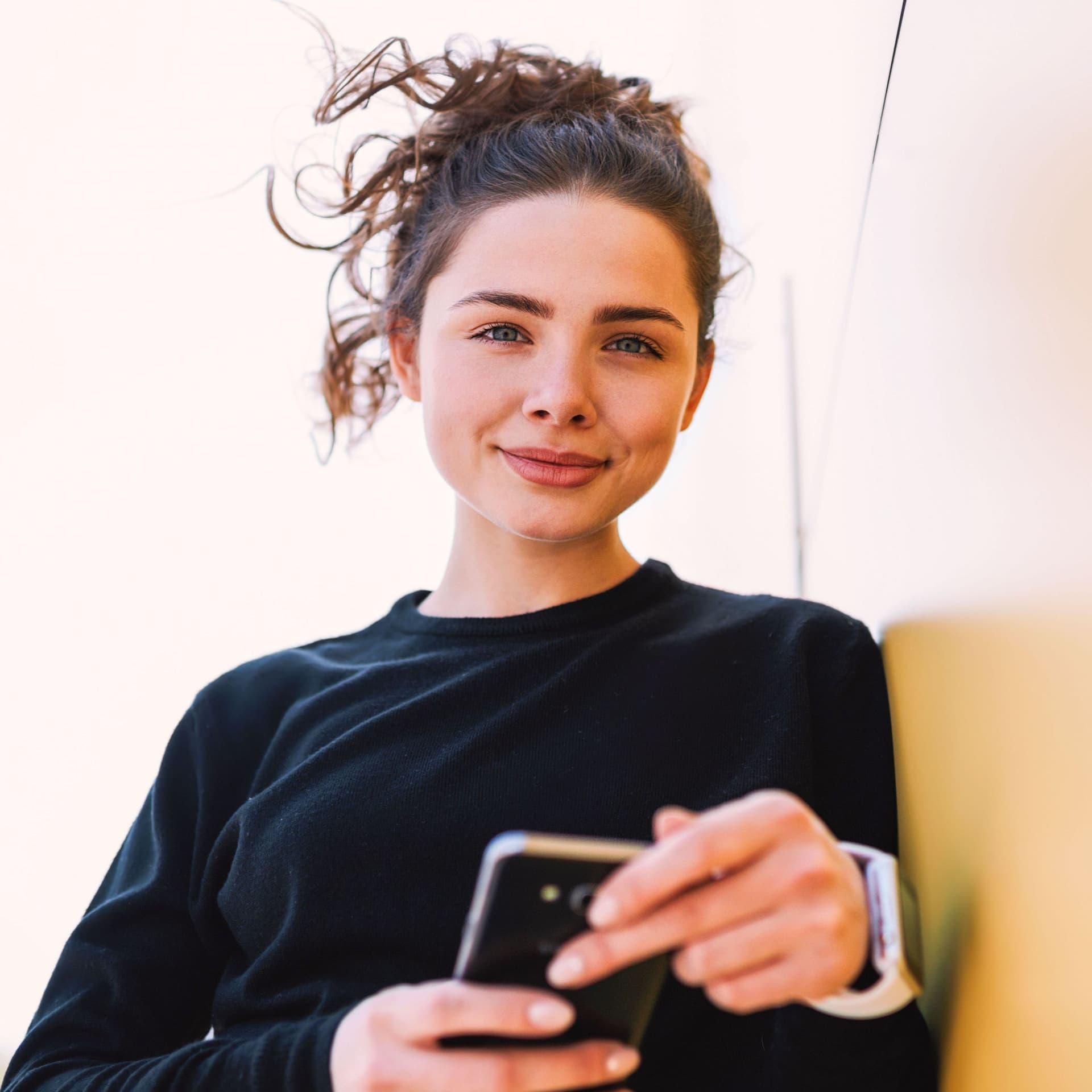 Junge Frau lehnt mit Handy an einer Wand und schaut in die Kamera.