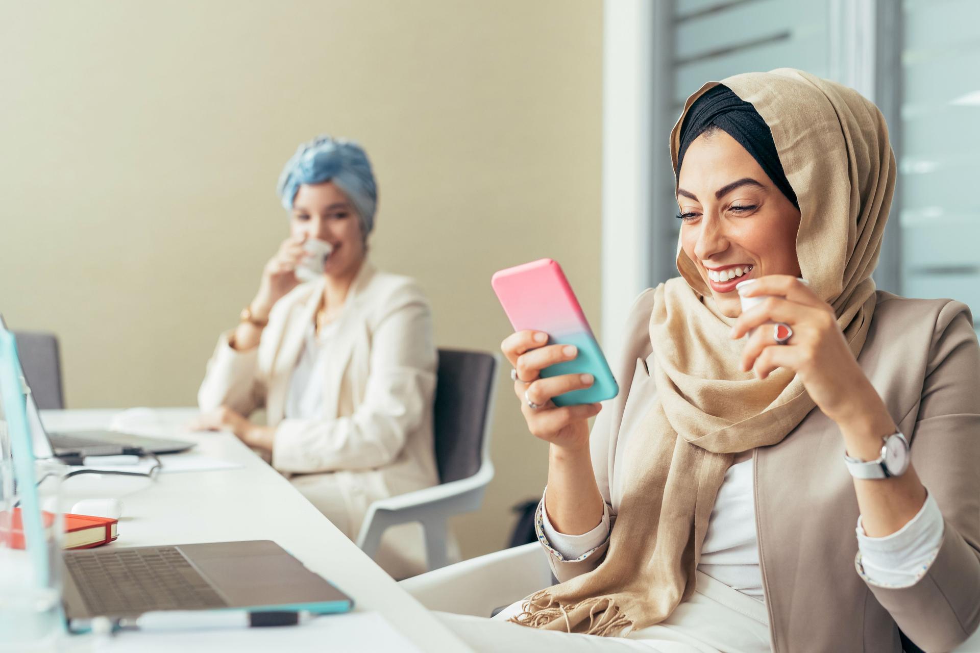 Frau mit Hijab blickt im Büro lachend auf ihr Smartphone.