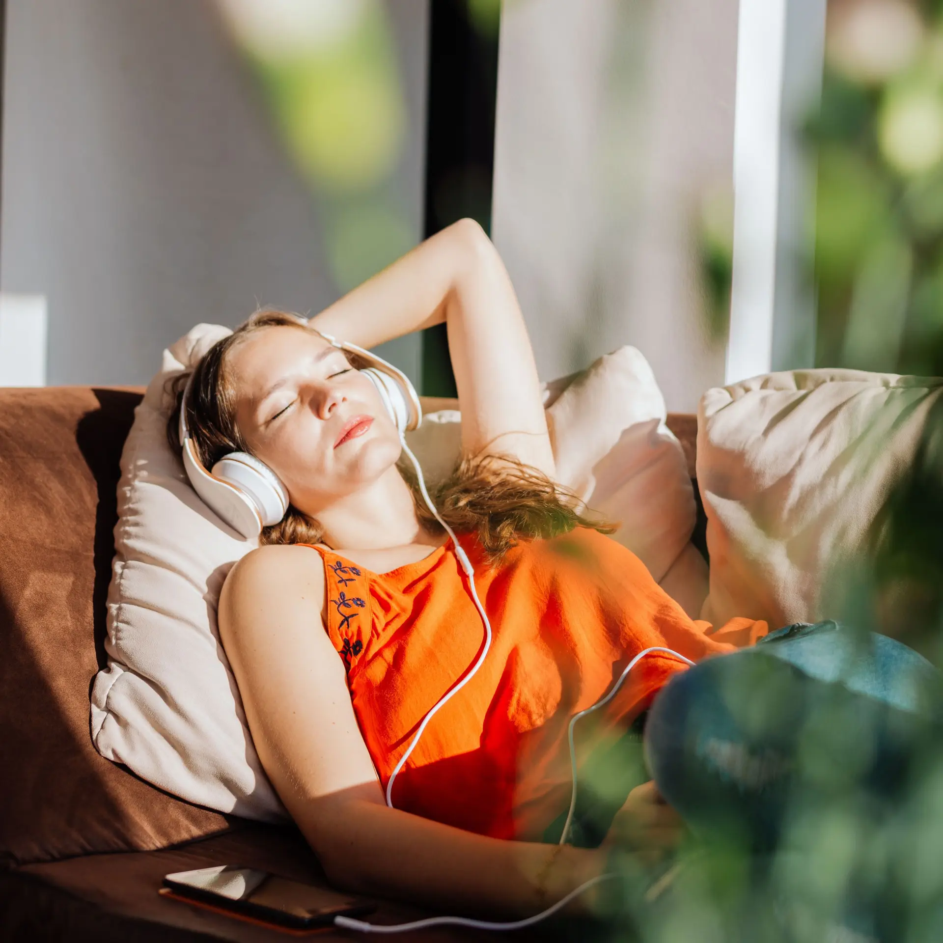 Eine junge Frau liegt entspannt auf der Couch. Die Sonne scheint ihr ins Gesicht. Sie hat Kopfhörer auf.
