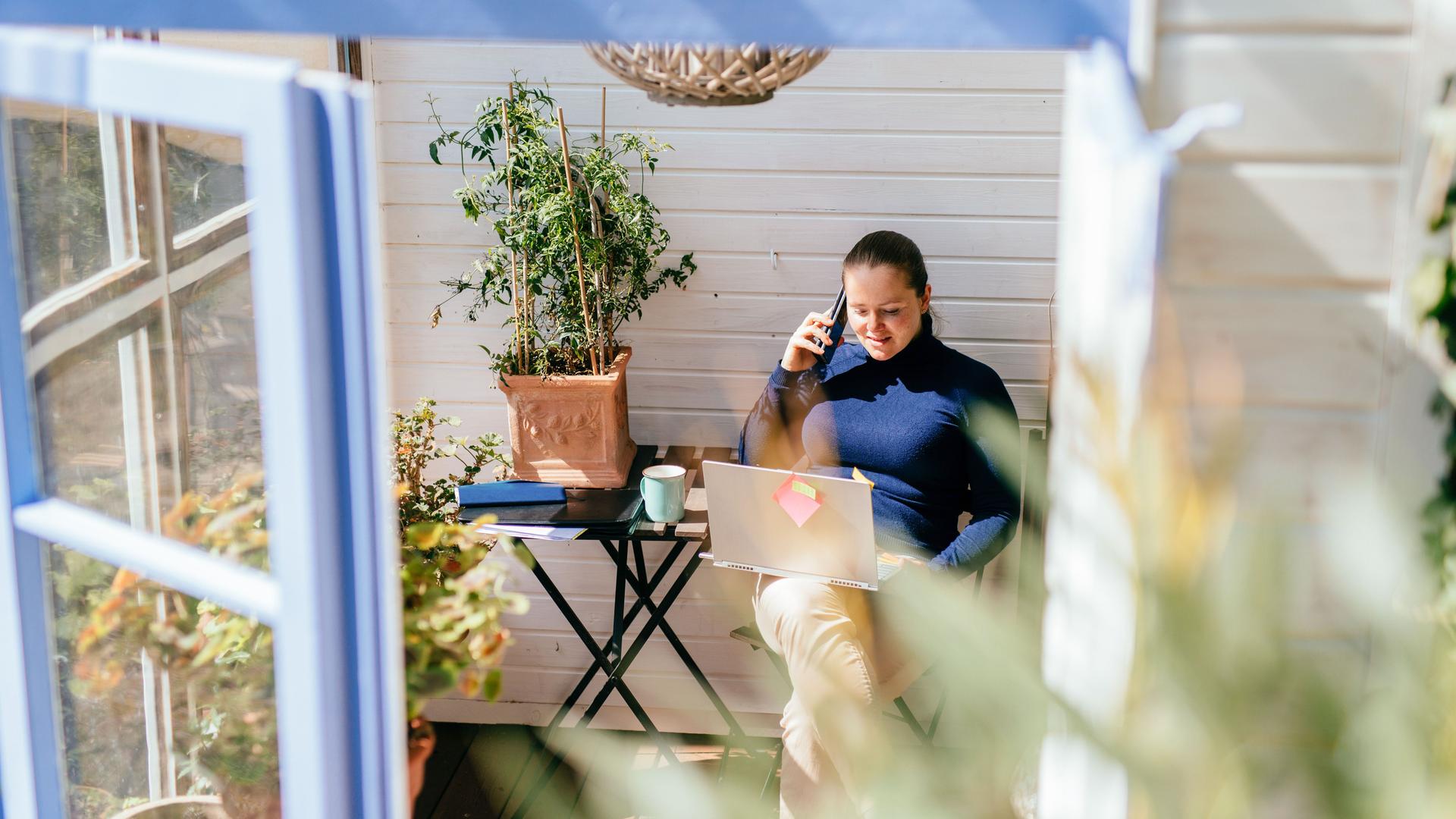 Frau sitzt mit Laptop auf dem Schoß und Mobiltelefon am Ohr in einem hellen Wintergarten. Blick von außen durch ein geöffnetes Fenster.