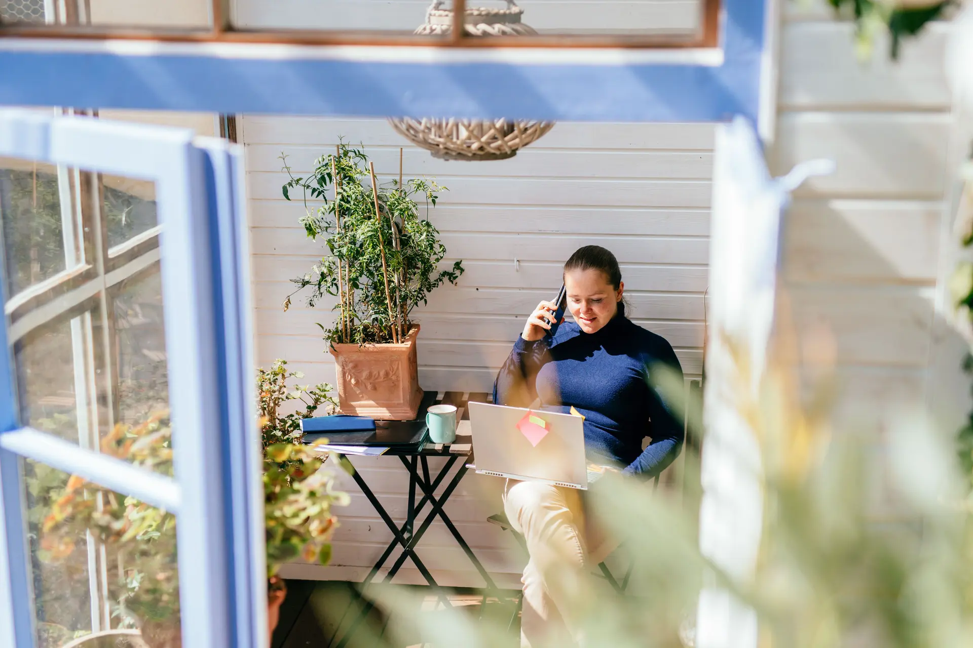Frau sitzt mit Laptop auf dem Schoß und Mobiltelefon am Ohr in einem hellen Wintergarten. Blick von außen durch ein geöffnetes Fenster.