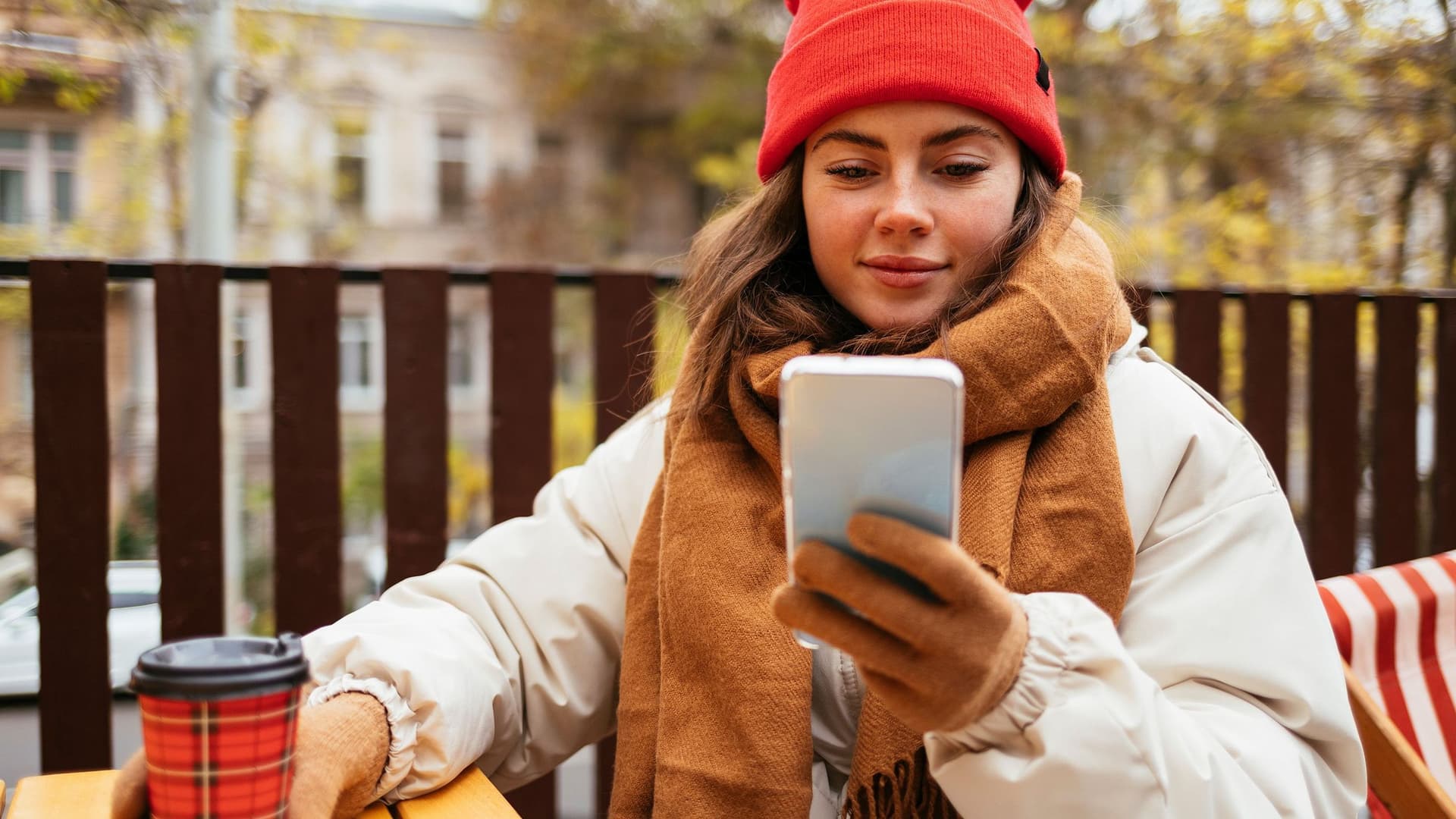 Eine Frau mit Mütze und Kaffee ind er Hand schaut auf ihr Smartphone