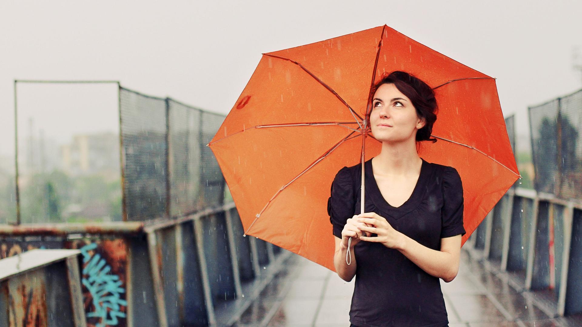 Junge Frau mit orangefarbenem Regenschirm im Regen auf einer Brücke.