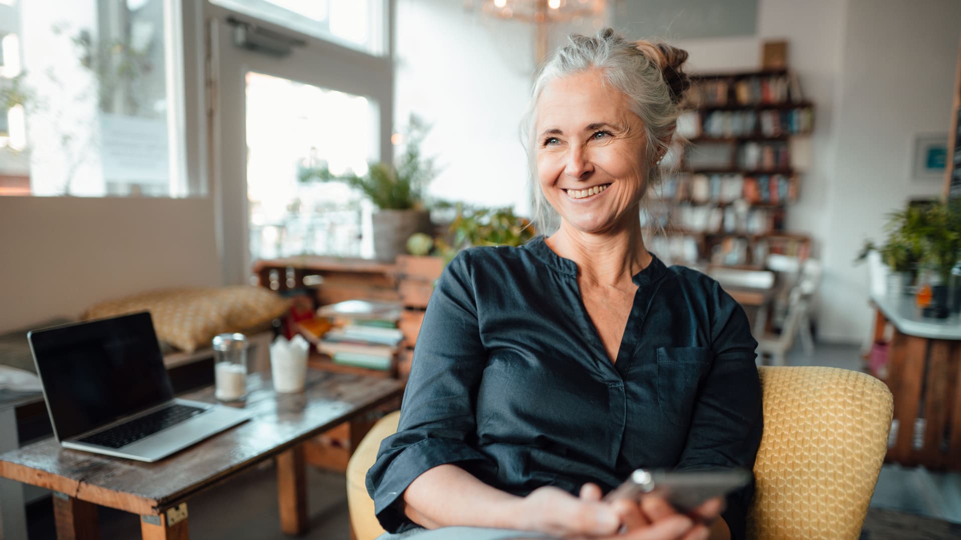 Lächelnde ältere Frau sitzt auf einem Sessel, blickt in die Ferne und hält ein Smartphone in der Hand.