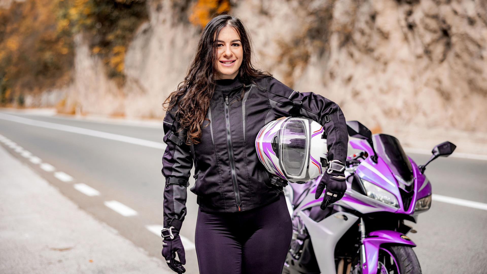 Junge Frau mit Helm unterm arm steht vor einem lilafarbenen Motorrad an einer Landstraße und lächelt in die Kamera.