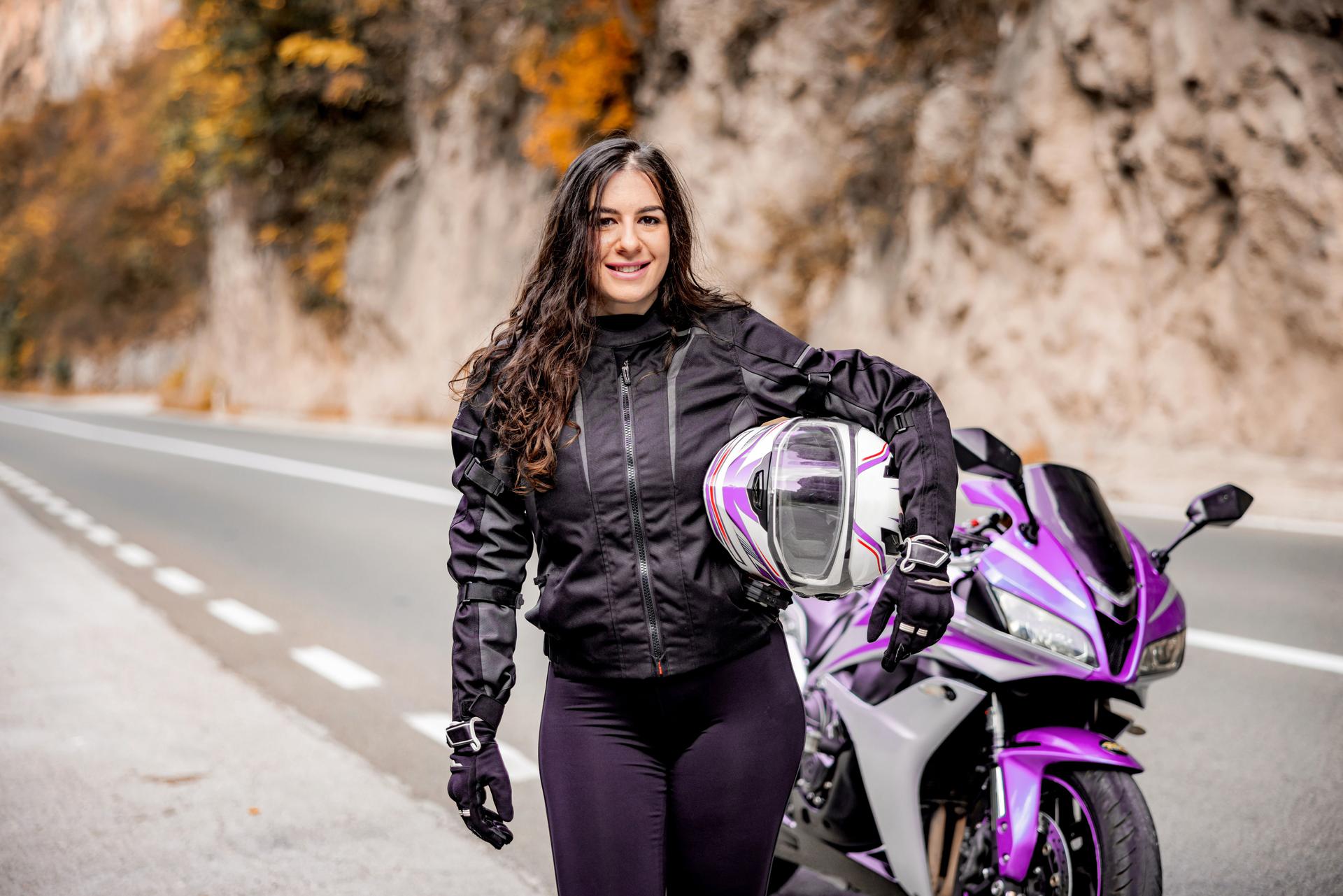 Junge Frau mit Helm unterm arm steht vor einem lilafarbenen Motorrad an einer Landstraße und lächelt in die Kamera.