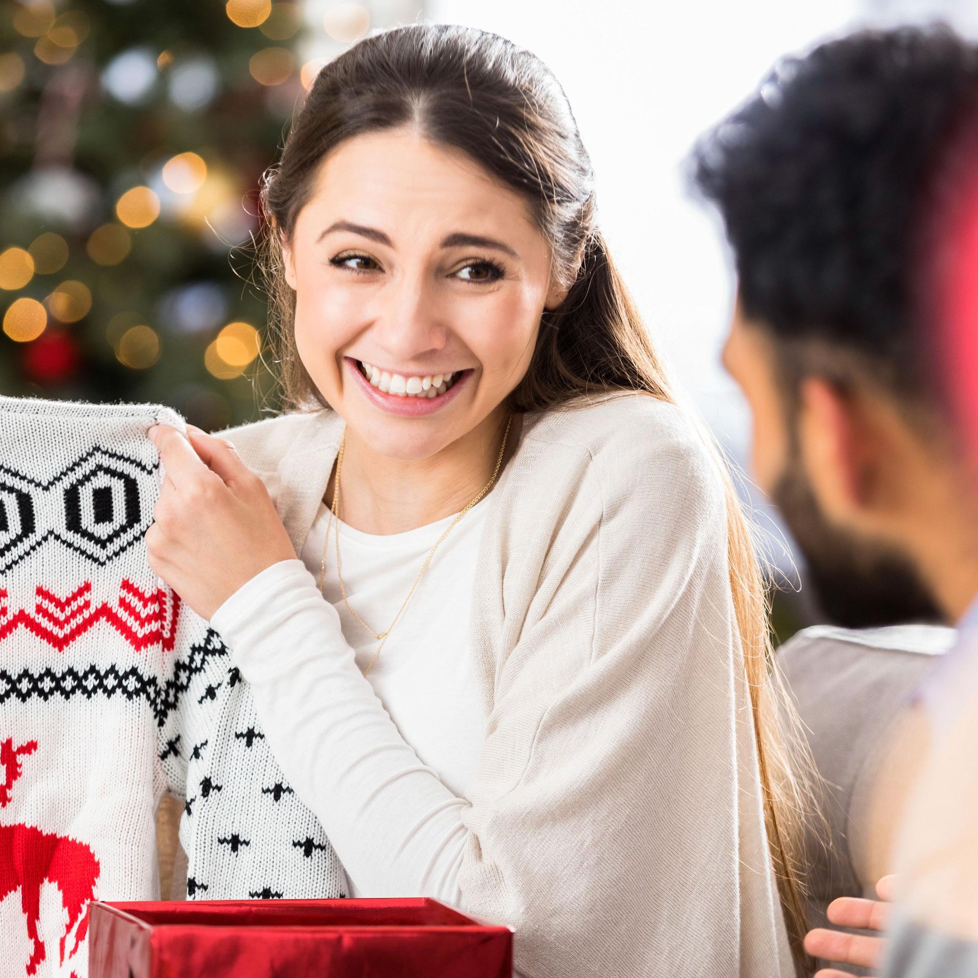 Eine junge Frau lächelt und verzieht das Gesicht, während sie einen Weihnachtspullover hochhält und ihn einem Mann zeigt.