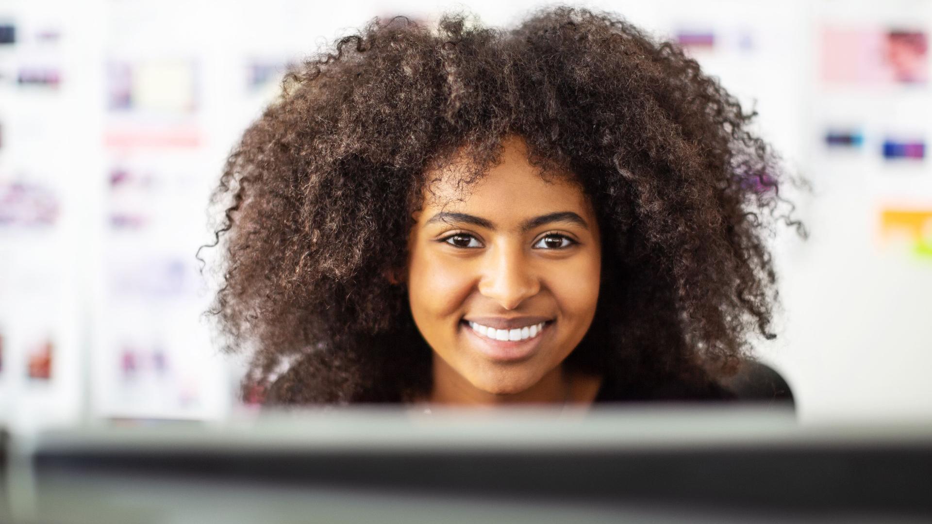 Porträt einer lachenden jungen Frau, die vor Ihrem Computer steht. Im Hintergrund sind Haftnotizen an einem Whiteboard zu sehen.