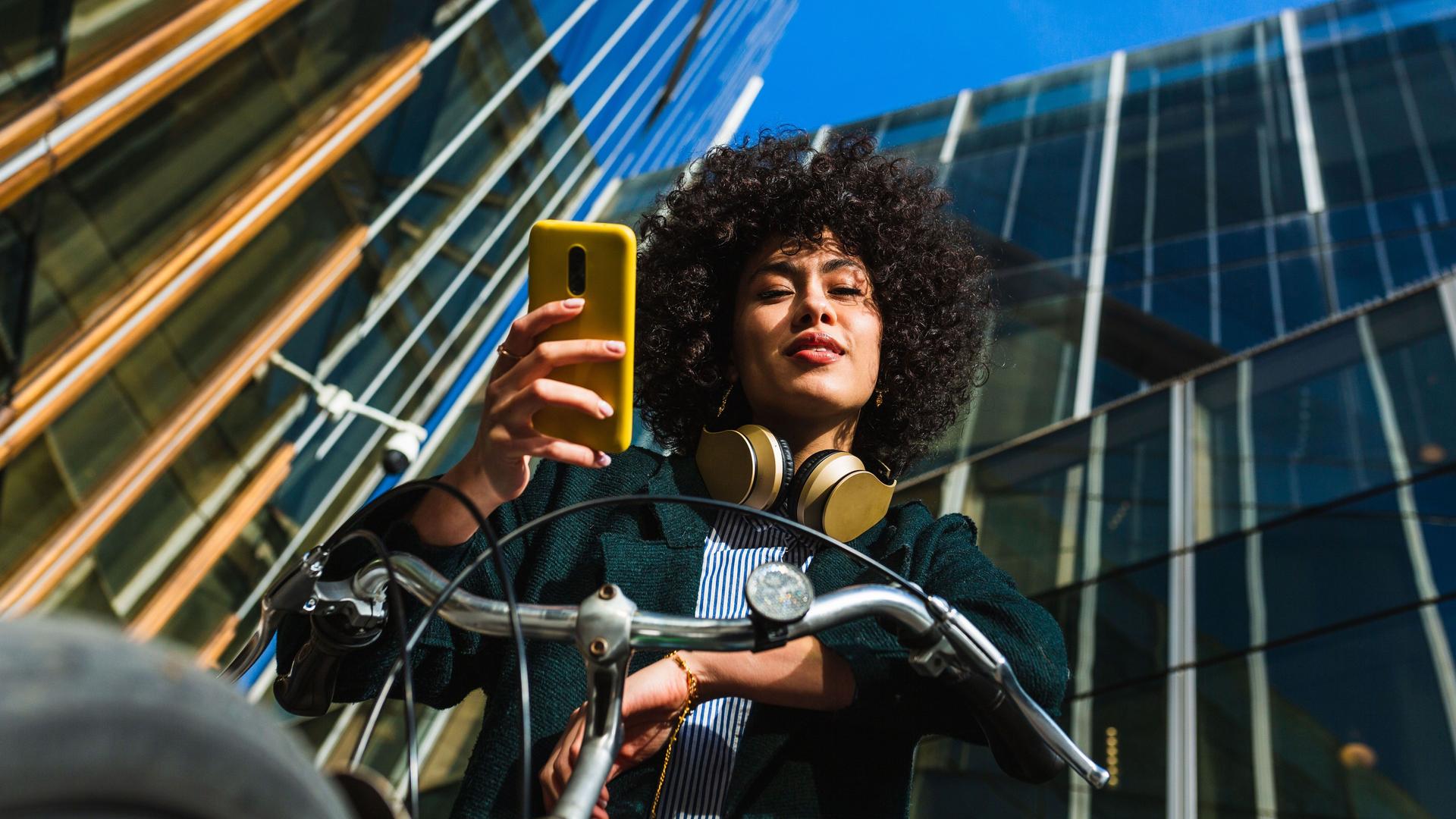 Eine Junge Frau auf einem Fahrrad aus der Froschperspektive hält ein Smartphone in der Hand. Im Hintergrund befinden dich verspiegelte Gebäude und ein blauer Himmel.