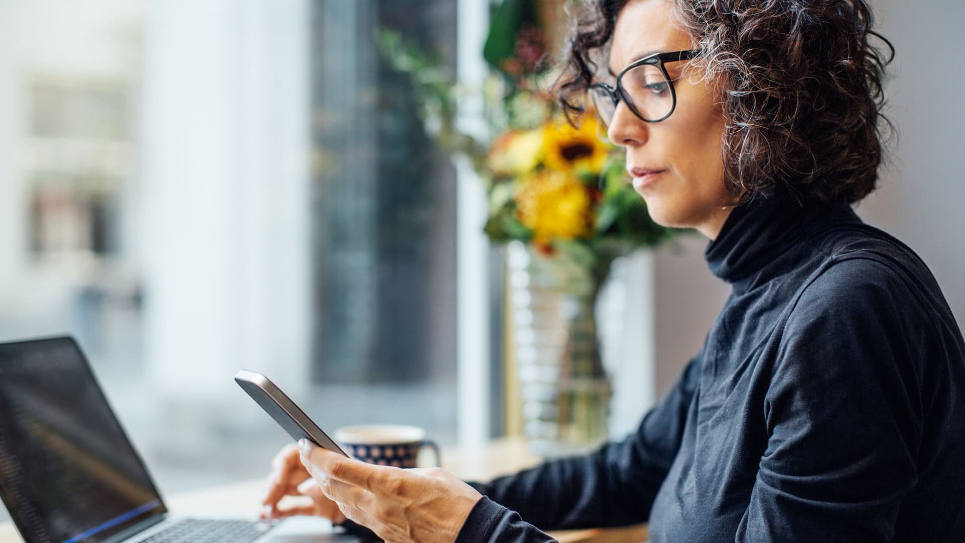 Frau sitzt in einem Cafe. Sie schaut auf ihr Smartphone. Vor ihr steht ein Laptop.