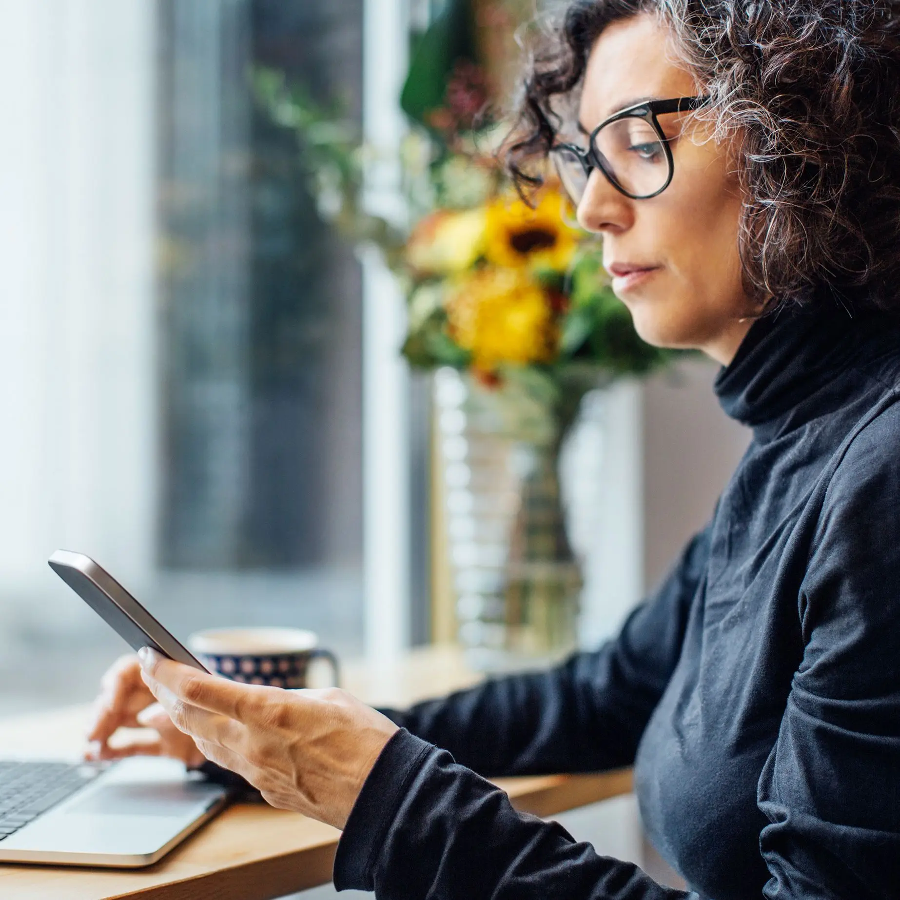 Frau sitzt in einem Cafe. Sie schaut auf ihr Smartphone. Vor ihr steht ein Laptop.