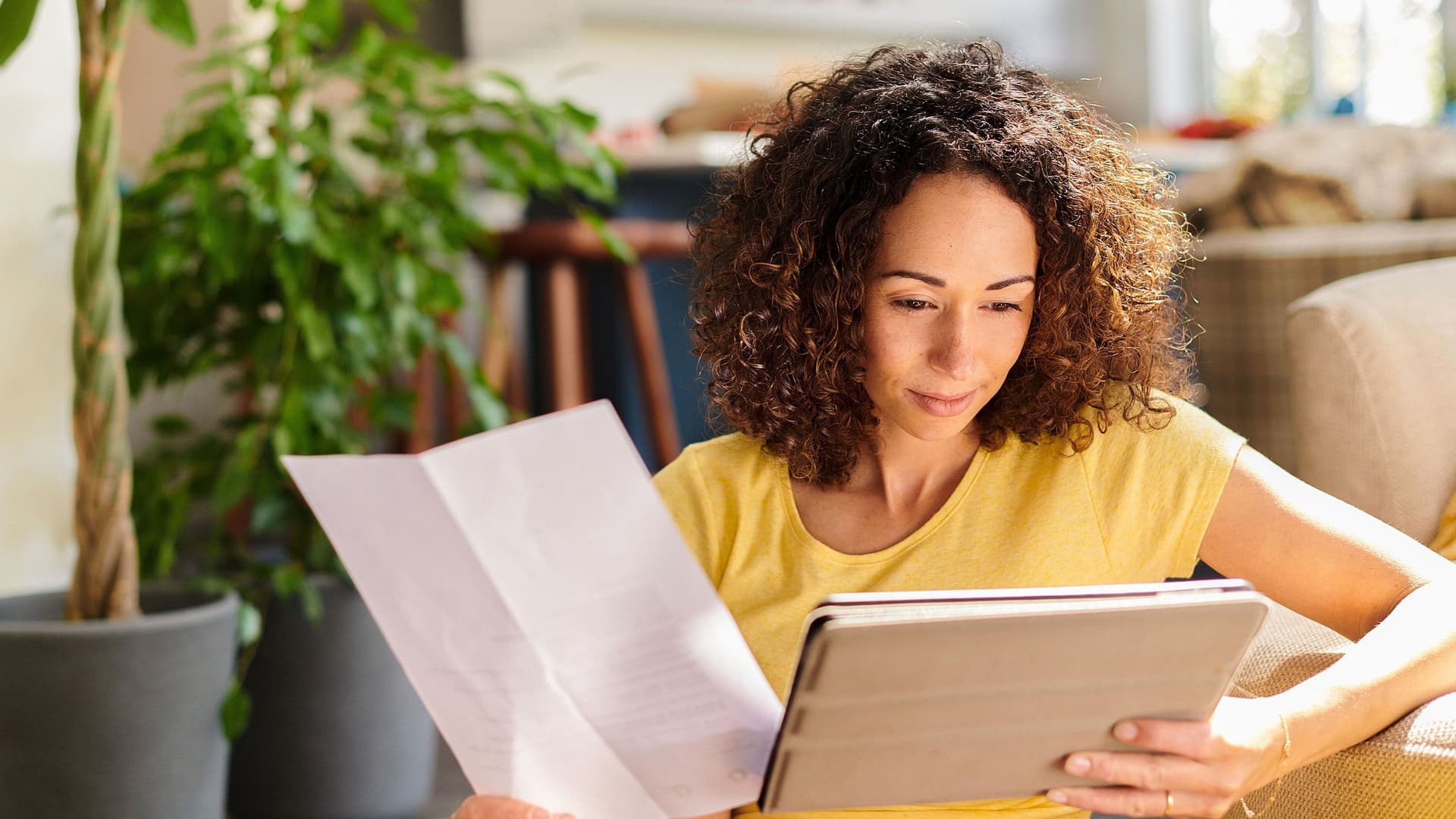 Eine Frau mit Dokumenten in der Hand schaut auf ein Tablet. Sie sitzt in ihrer sonnigen Wohnung.