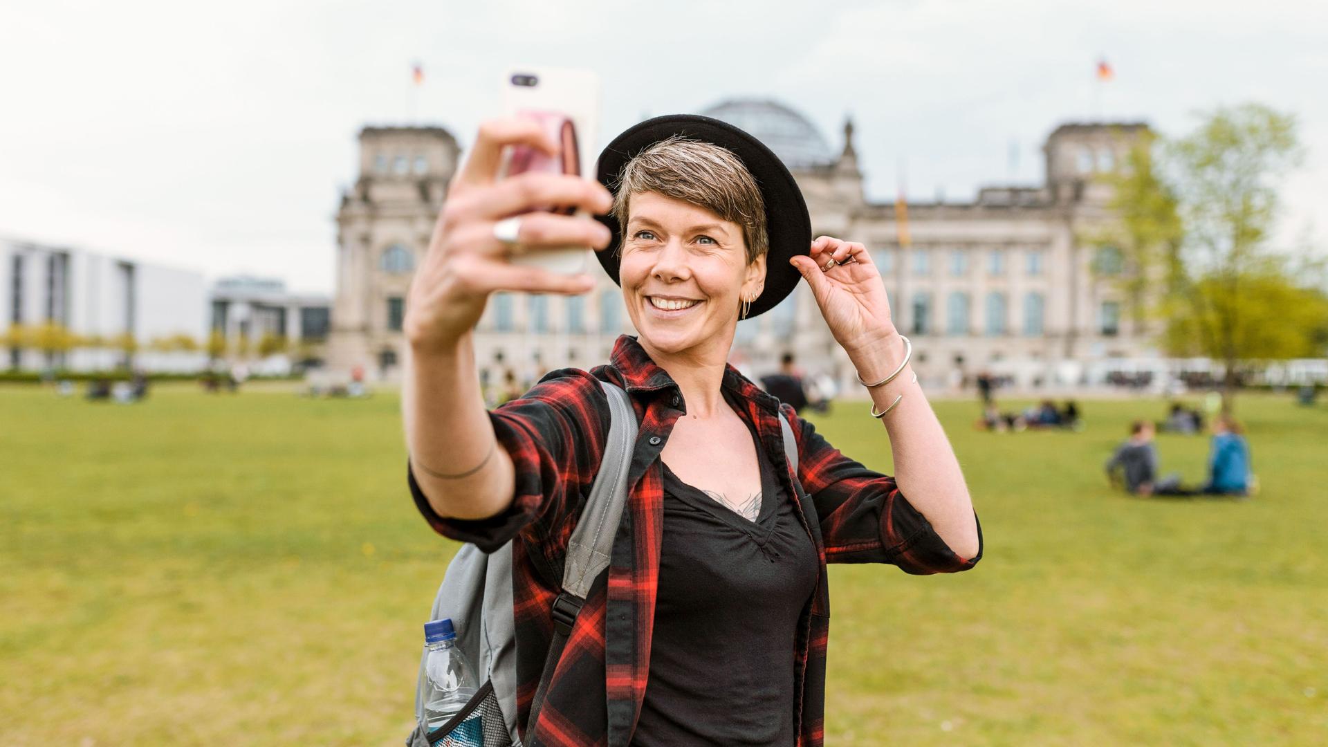 Eine Frau mittleren Alters mit Hut und Rucksack steht vor dem Bundestag. Sie macht ein Selfie und lächelt in Richtung Smartphone.