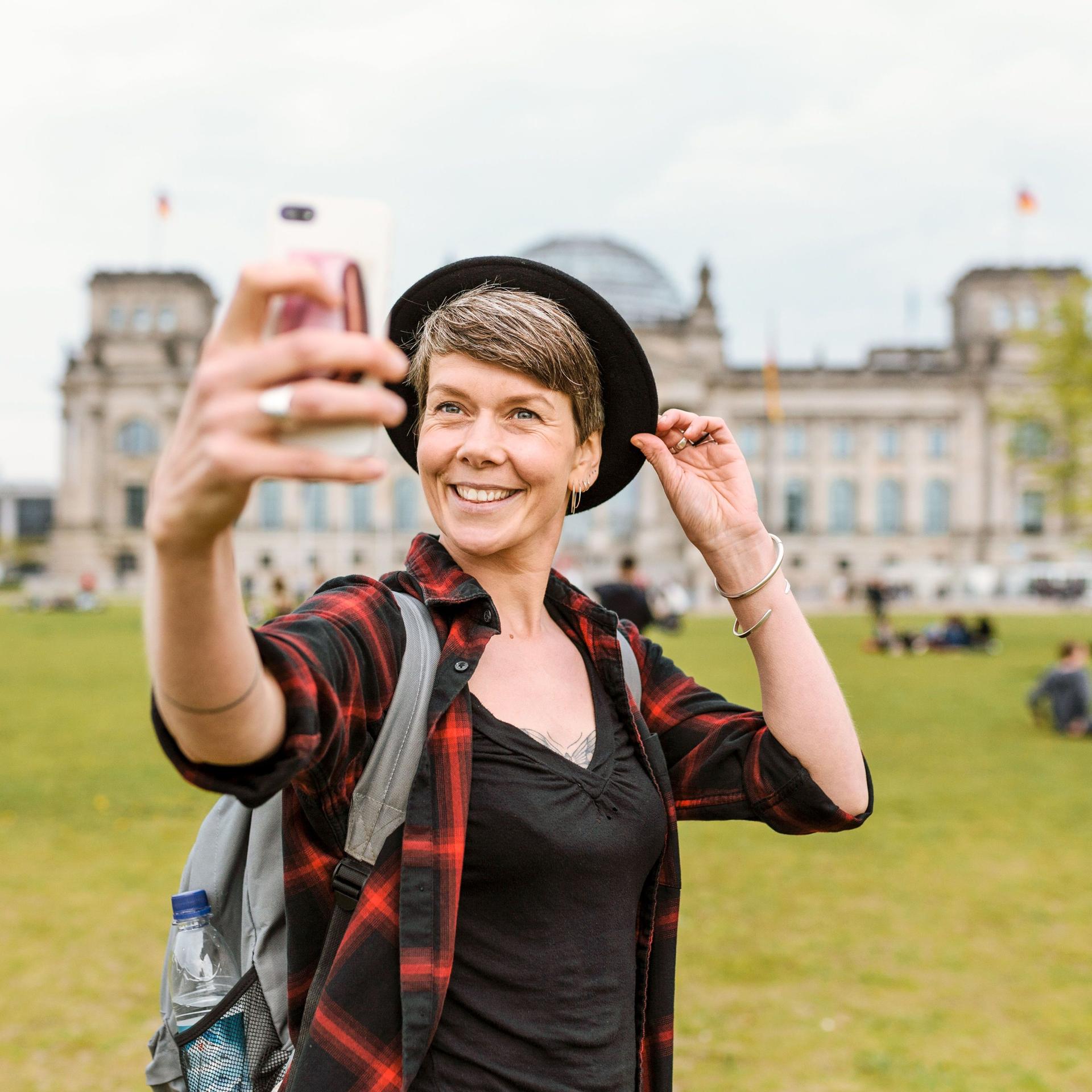 Eine Frau mittleren Alters mit Hut und Rucksack steht vor dem Bundestag. Sie macht ein Selfie und lächelt in Richtung Smartphone.