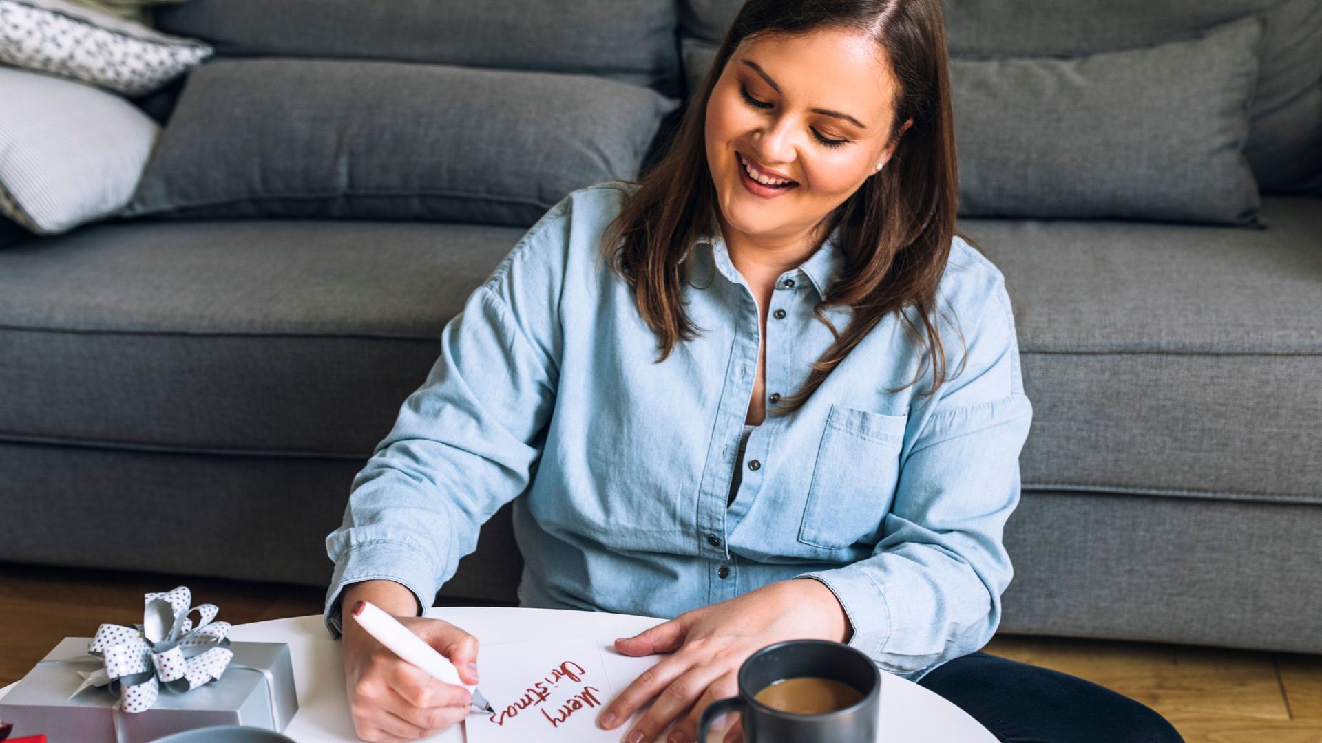 Eine lächelnde Frau, die im Wohnzimmer auf dem Boden sitzt und eine Weihnachtskarte auf dem Couchtisch schreibt. Auf dem Tische stehen Kekse und ein Kaffee.