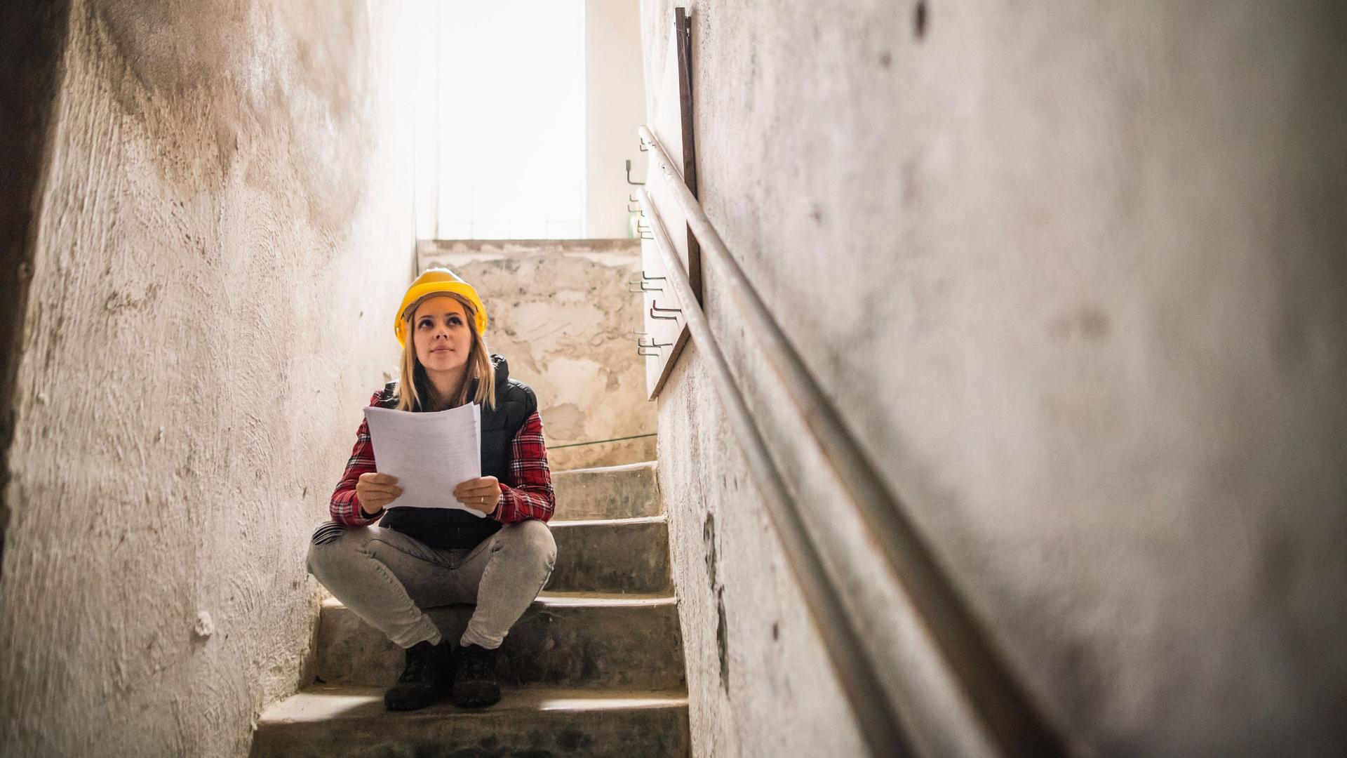 Junge Frau mit einem gelben Helm sitzt auf einer Steintreppe auf einer Baustelle. Sie hält Pläne in den Händen.