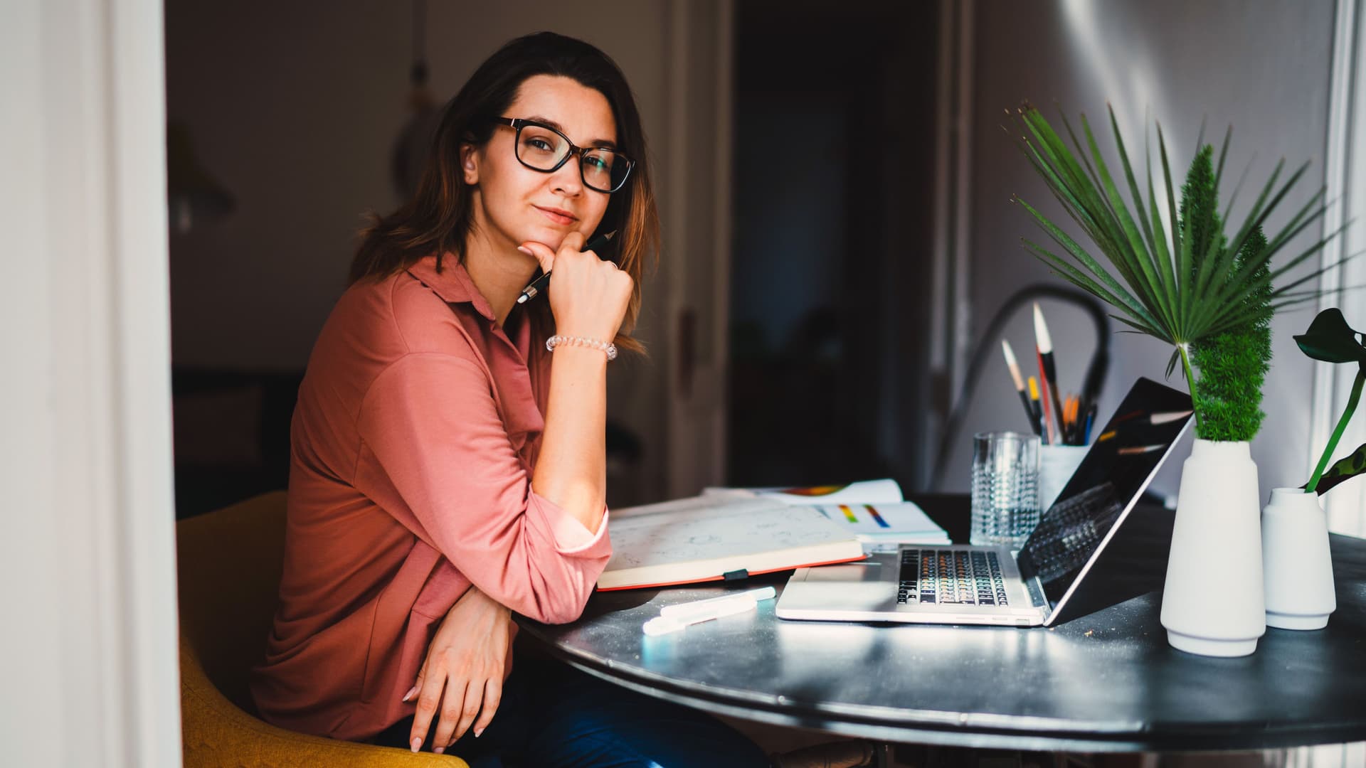 Junge Frau mit Brille lächelt in die Kamera. Sie sitzt an einem runden Tisch vor einem Laptop und aufgeschlagenen Büchern.