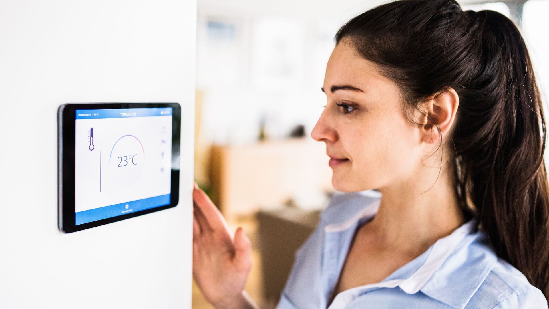 Eine Frau schaut auf ein Tablet an der Wand mit Smart Home-Bildschirm. Das Display zeigt die Temperatur 23°C.