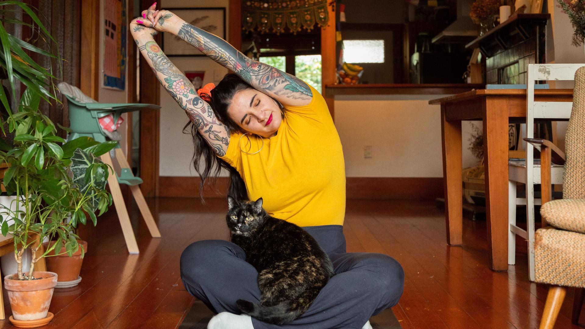 Tätowierte junge Frau streckt sich zu Hause mit Katze auf dem Schoß.