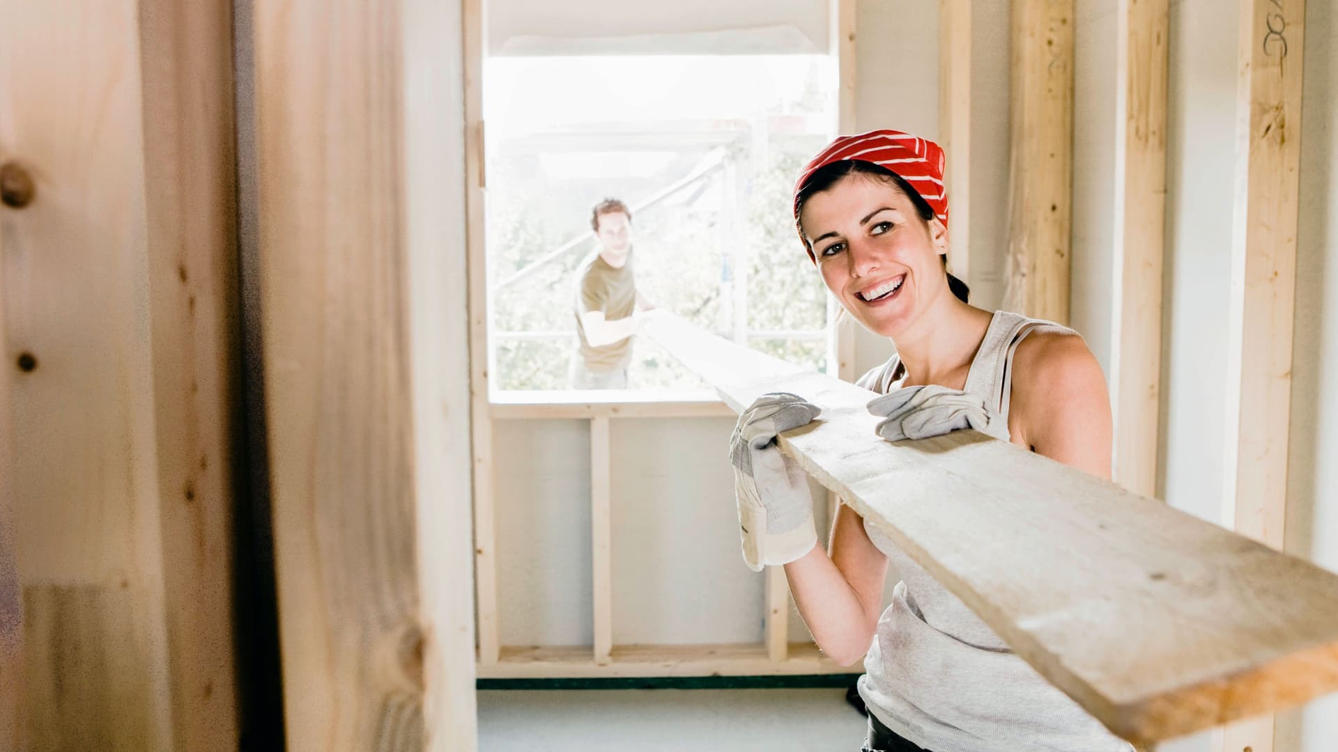 Eine Frau trägt ein Holzbrett durch das Fenster eines Rohbaus. Im Hintergrund ist draußen ein Mann zu sehen, der ebenfalls das Brett trägt.