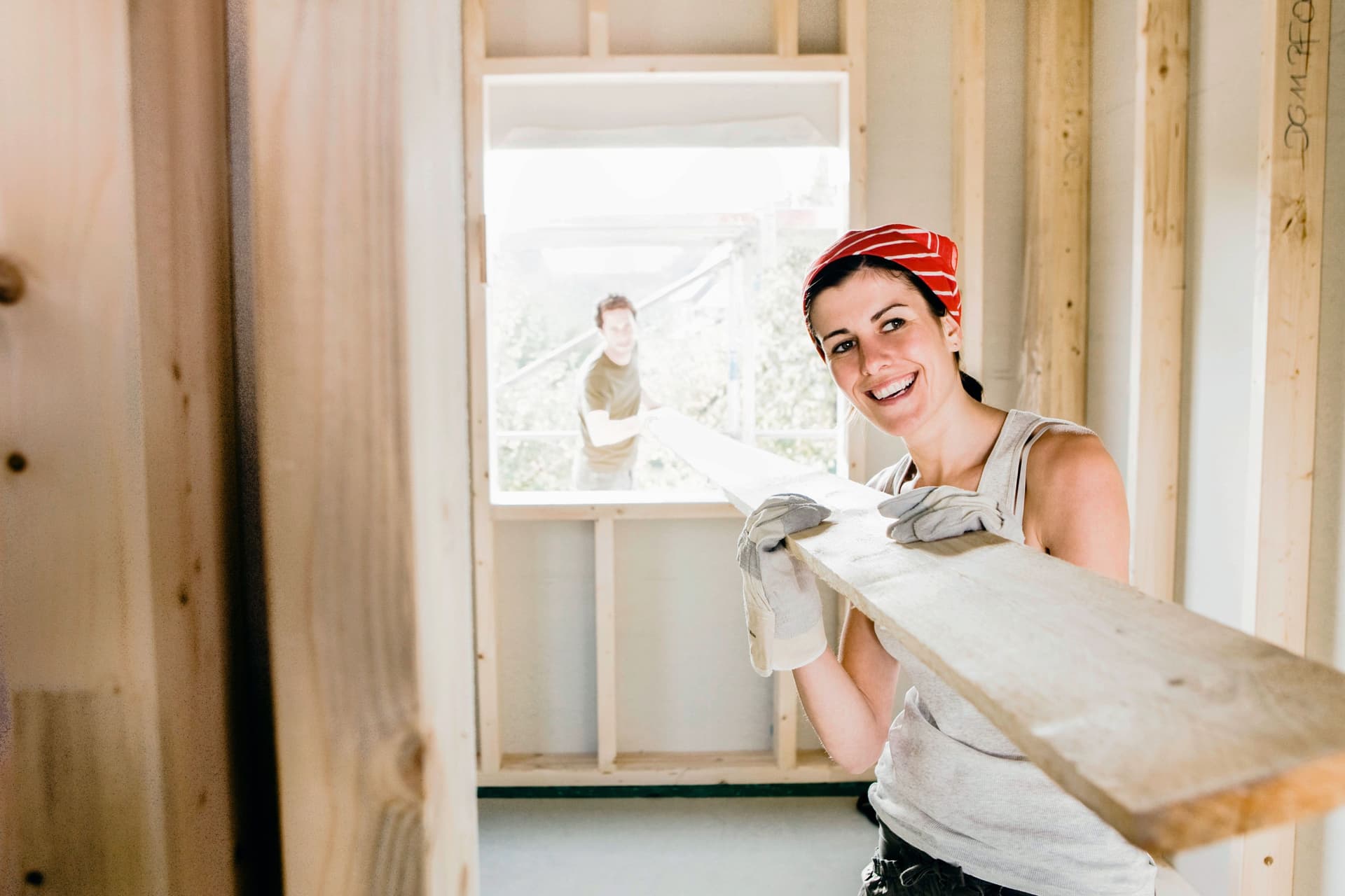 Eine Frau trägt ein Holzbrett durch das Fenster eines Rohbaus. Im Hintergrund ist draußen ein Mann zu sehen, der ebenfalls das Brett trägt.