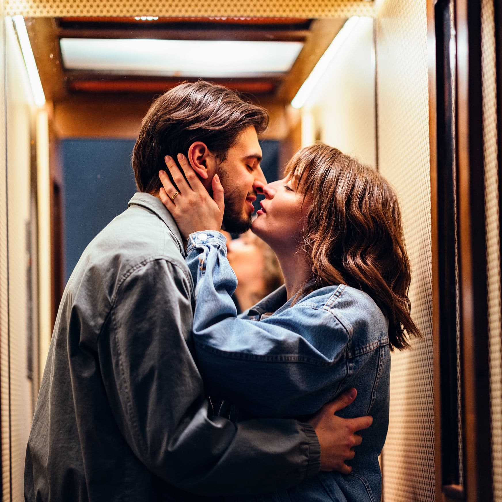 Ein junges Paar küsst sich in einem Aufzug mit warmem Licht, beide tragen Jeansjacken, sie hält sein Gesicht, er umarmt sie an der Taille.