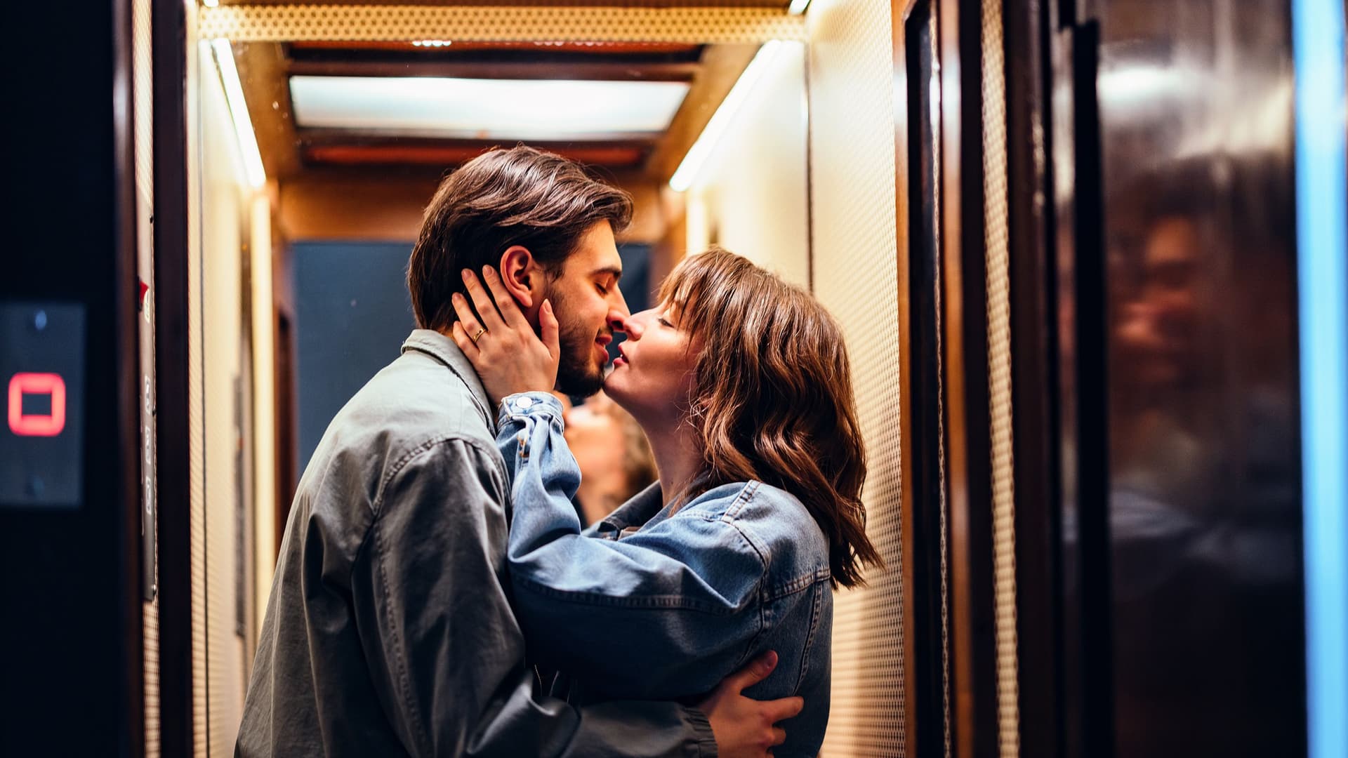 Ein junges Paar küsst sich in einem Aufzug mit warmem Licht, beide tragen Jeansjacken, sie hält sein Gesicht, er umarmt sie an der Taille.