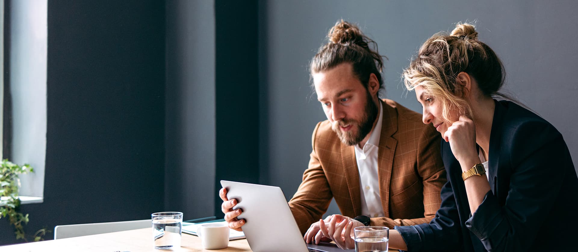Zwei Personen, eine Frau und ein Mann, sitzen an einem Tisch und schauen gemeinsam auf einen Laptop-Bildschirm. Auf dem Tisch liegen Unterlagen, daneben stehen zwei Wassergläser und eine Tasse.