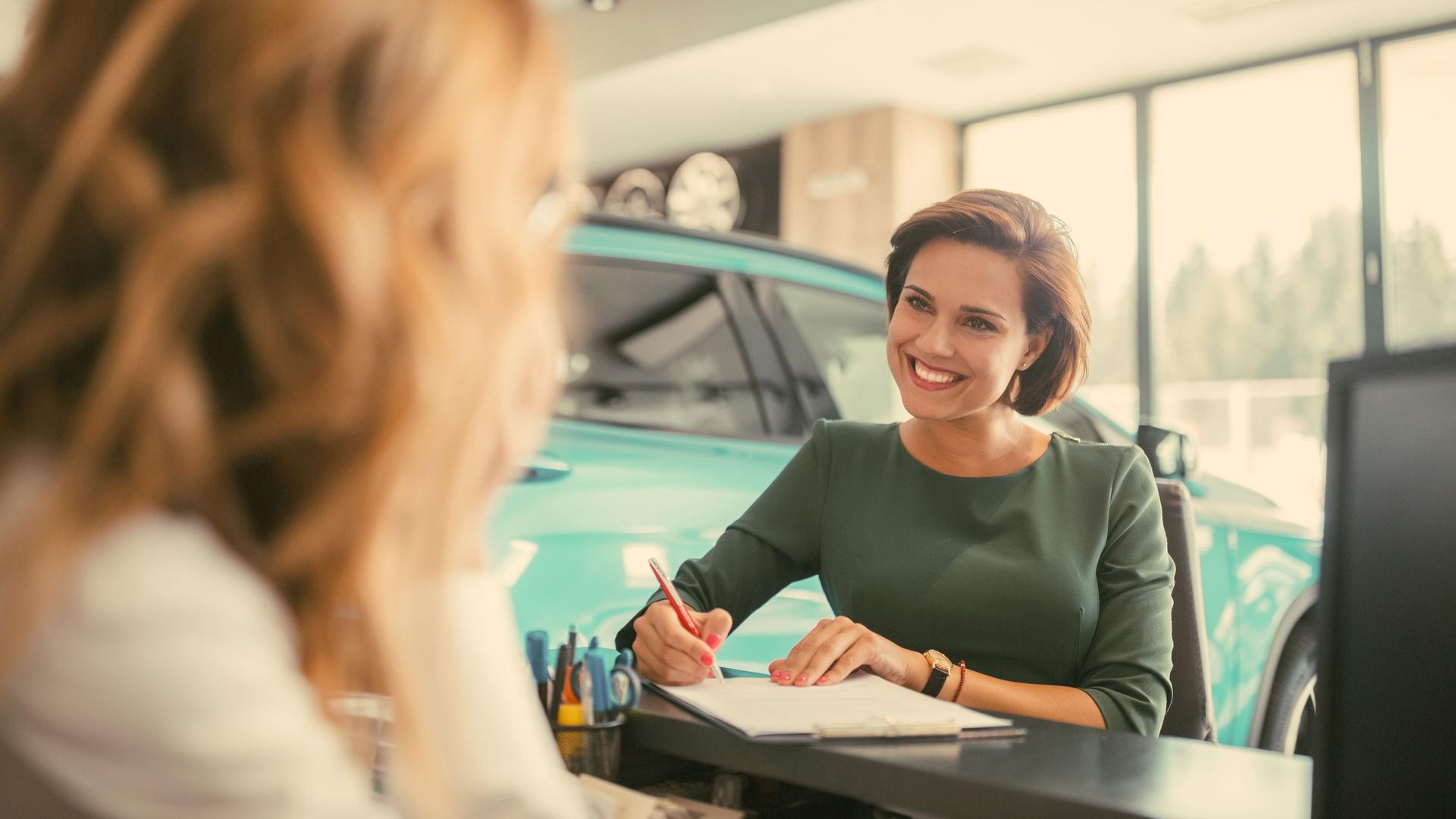 Zwei Frauen in einem Autohaus. Eine Frau unterschreibt lächelnd ein Dokument. 