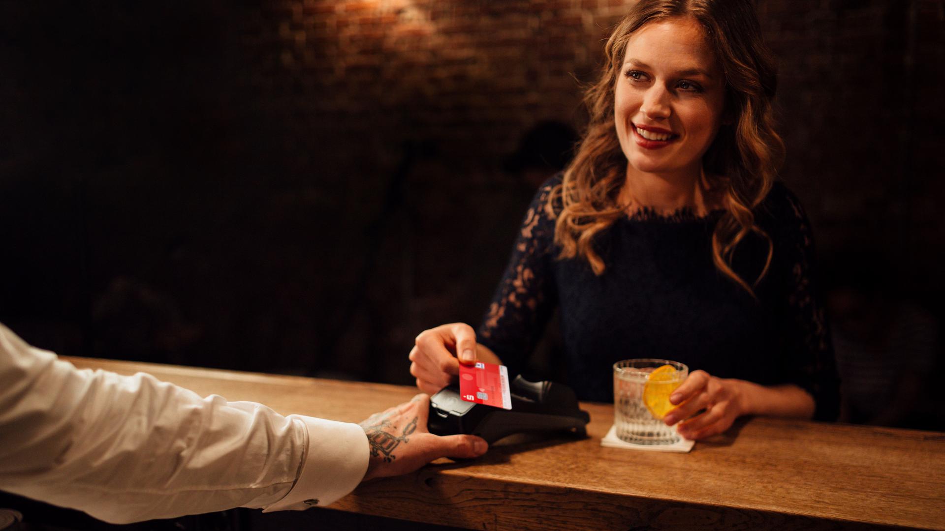 Festlich gekleidete junge Frau in einer Bar zahlt kontaktlos mit ihrer Sparkassenkarte. Sie lächelt den Barkeeper dabei an.