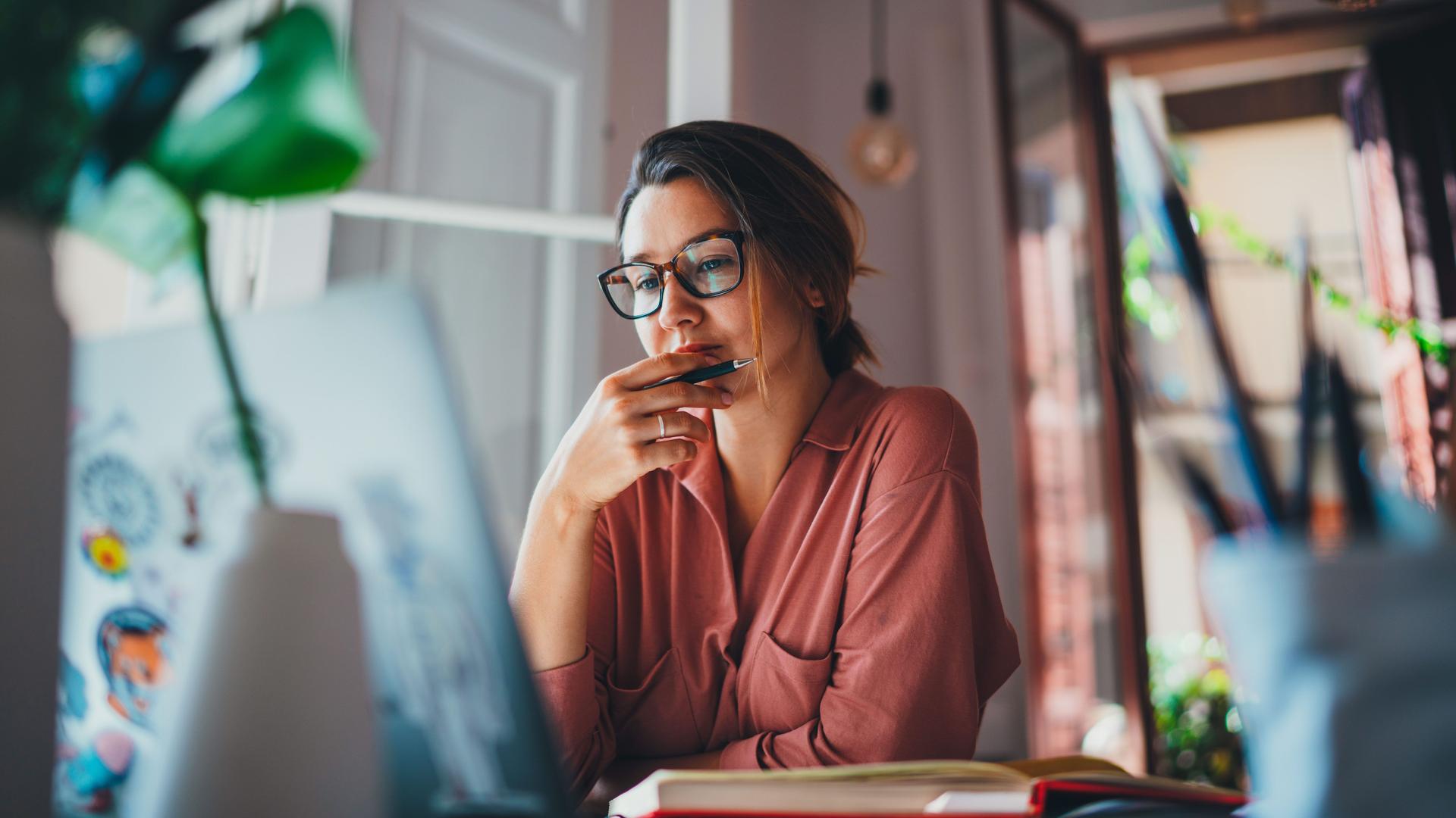 Junge Frau mit Brille, die zu Hause über etwas nachdenkt, während sie vor einem geöffneten Laptop sitzt.
