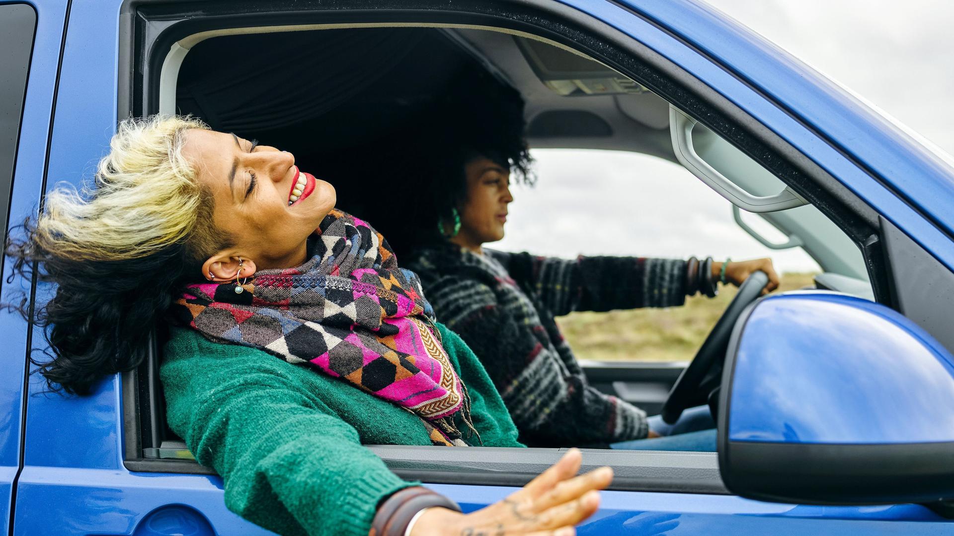Zwei Frauen fahren in einem blauen Van. Das Fenster beim Beifahrersitz ist offen und die Frau streckt lachend ihren Kopf und ihre Hand aus dem Fenster.