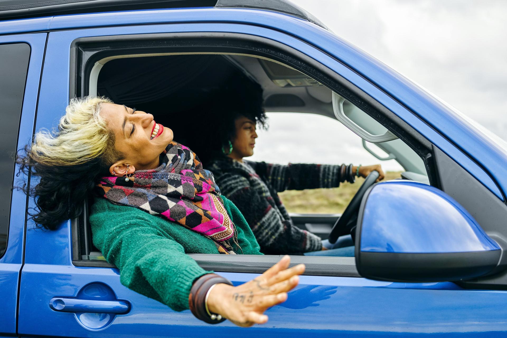 Zwei Frauen fahren in einem blauen Van. Das Fenster beim Beifahrersitz ist offen und die Frau streckt lachend ihren Kopf und ihre Hand aus dem Fenster.