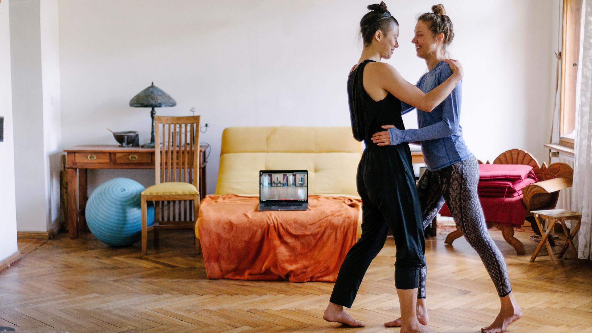 Zwei Frauen tanzen zusammen Tango in ihrer Altbauwohnung. Im Hintergrund steht ein Laptop, auf dem ein Online-Tanzkurs zu sehen ist.