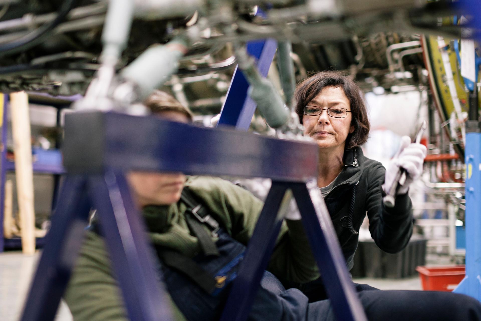 Zwei Mechanikerinnen arbeiten an einer Maschine in einem Flugzeughangar. Sie sind umgeben von Motoren und Drähten.
