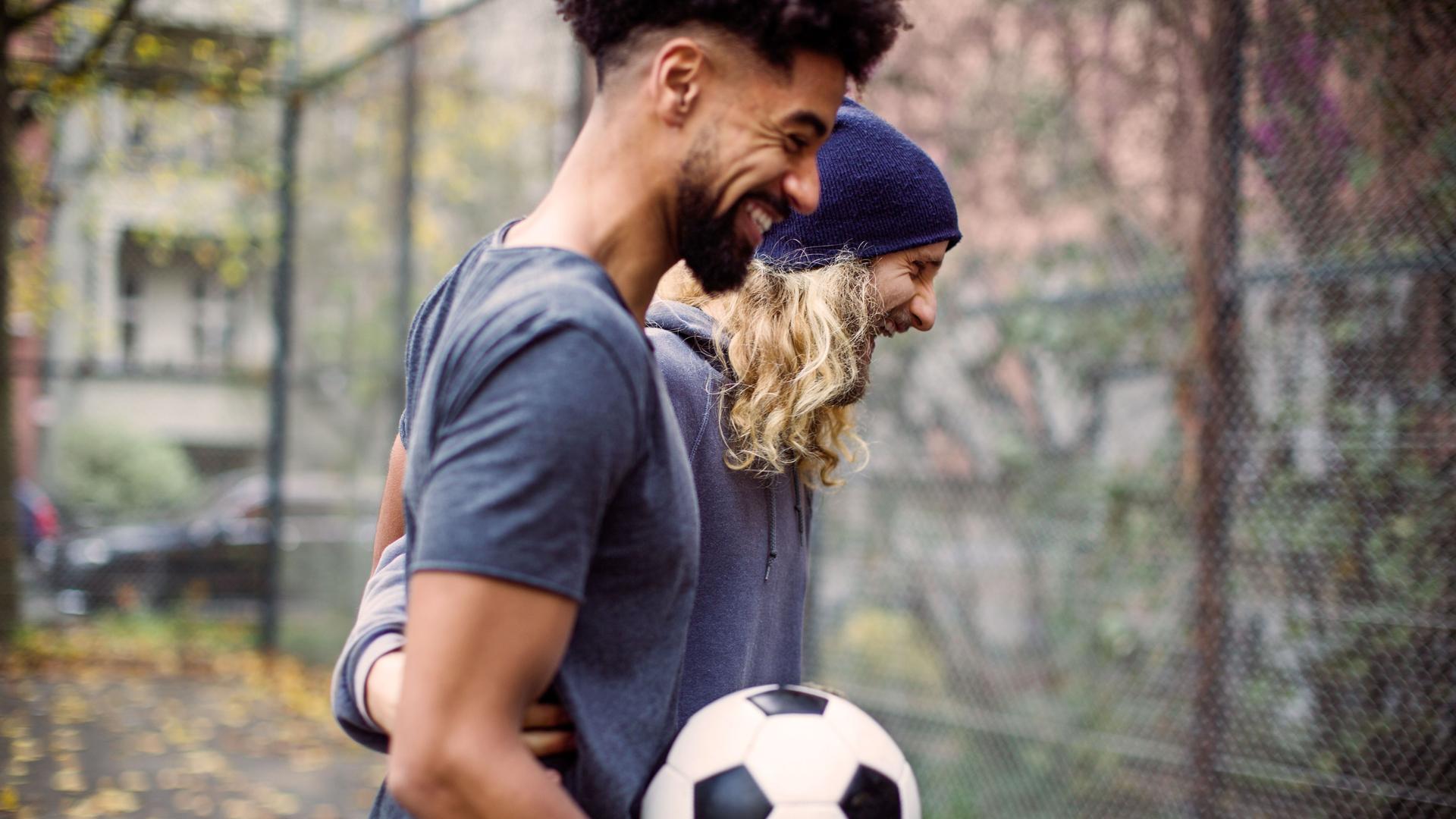 Zwei lachende Fußballspieler mit Ball im Gespräch auf einem asphaltierten Fußballplatz. Sie umarmen sich und lachen ausgelassen.