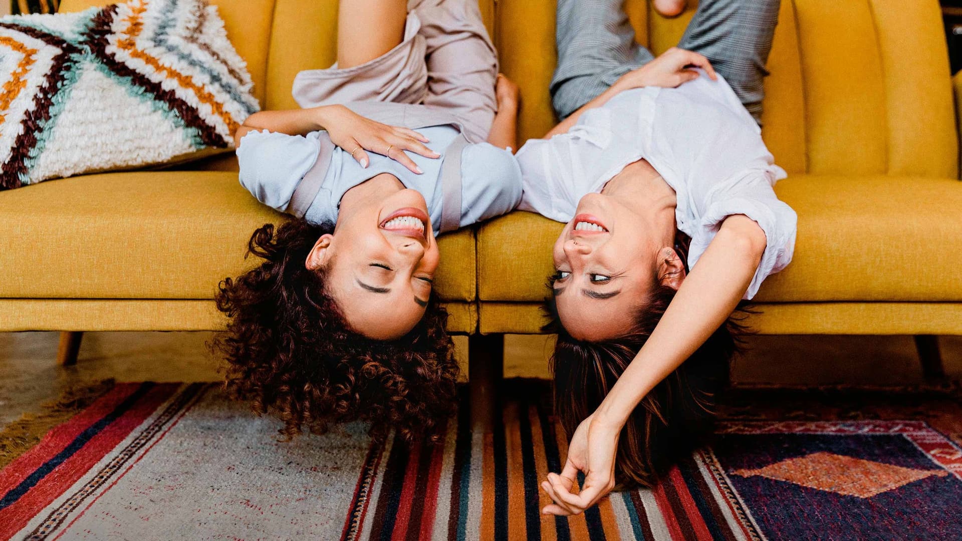 Zwei junge Frauen liegen kopfüber ausgelassen und lachend auf einem gelben Sofa in einem Wohnzimmer.