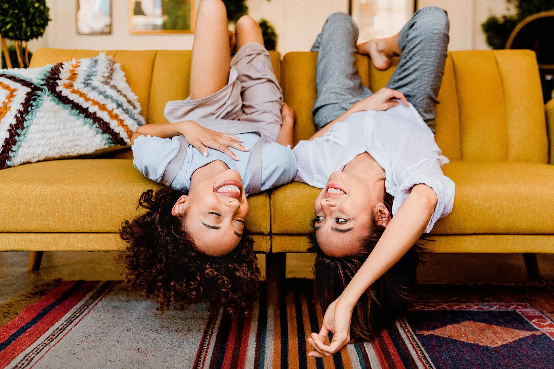 Zwei junge Frauen liegen kopfüber ausgelassen und lachend auf einem gelben Sofa in einem Wohnzimmer.