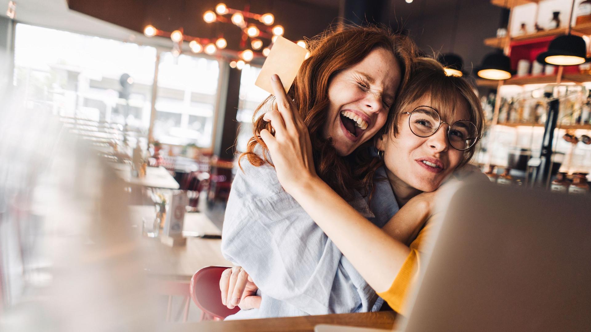 Zwei Freundinnen umarmen sich euphorisch. Sie sitzen in einem Cafe vor einem Laptop. Eine der jungen Frauen hält eine Bankkarte hoch.