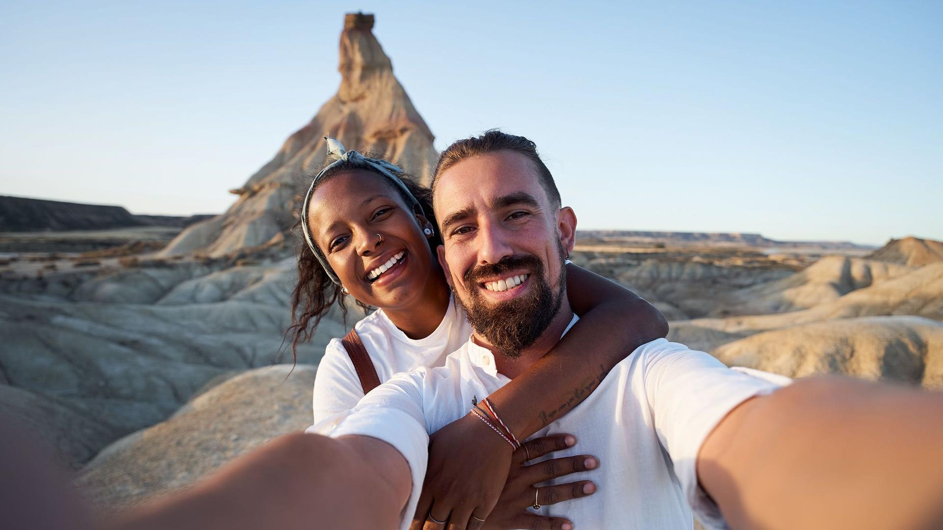 Fröhliches Selfie eines Paares in einer steinigen Wüstenlandschaft mit einem Felsberg im Hintergrund.