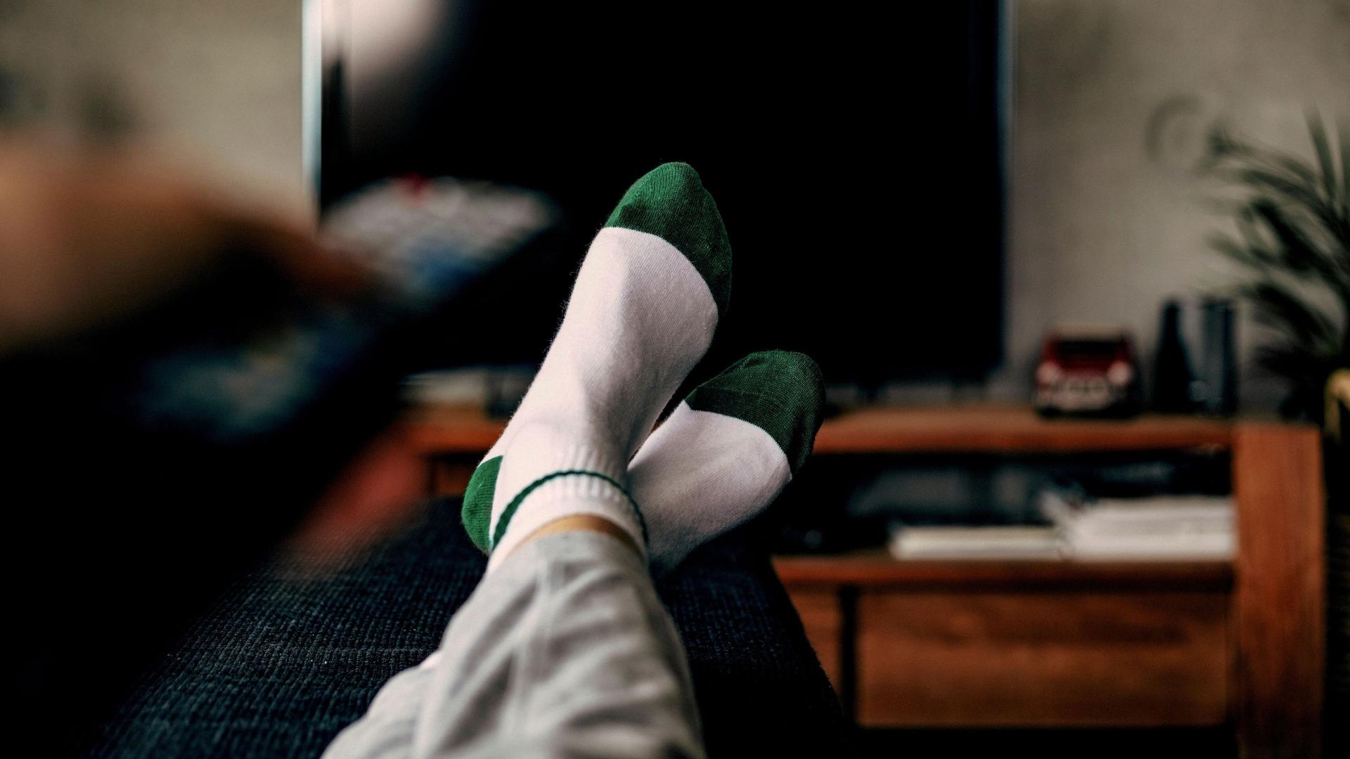 Eine Person sitzt mit hochgelegten Füßen auf der Couch und hält eine Fernbedienung in der Hand, die auf den Fernseher gerichtet ist.