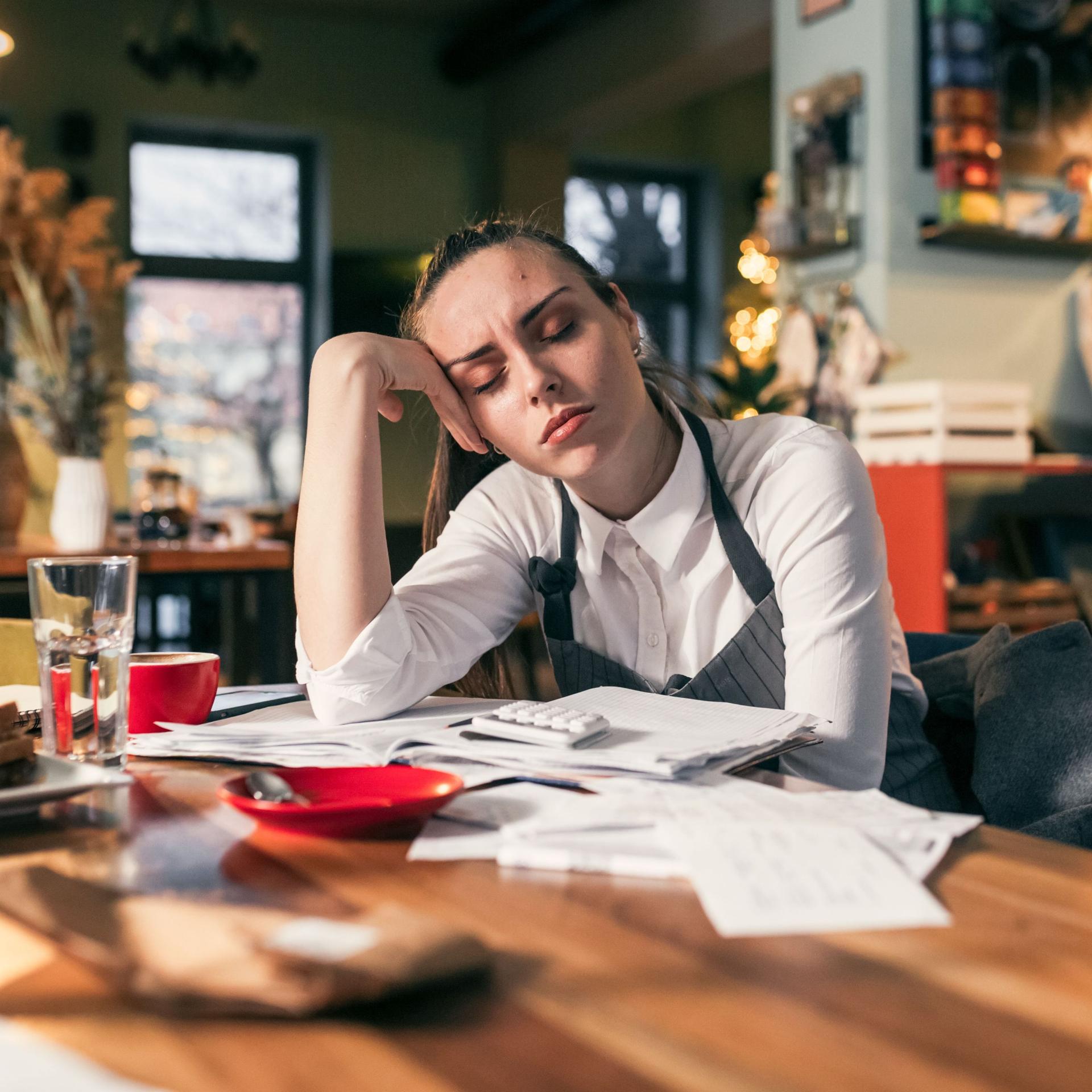 Eine traurig aussehende junge Frau aus der Gastronomie sitzt mit geschlossenen Augen über Rechnungen an einem Tisch in gemütlicher Atmosphäre. 