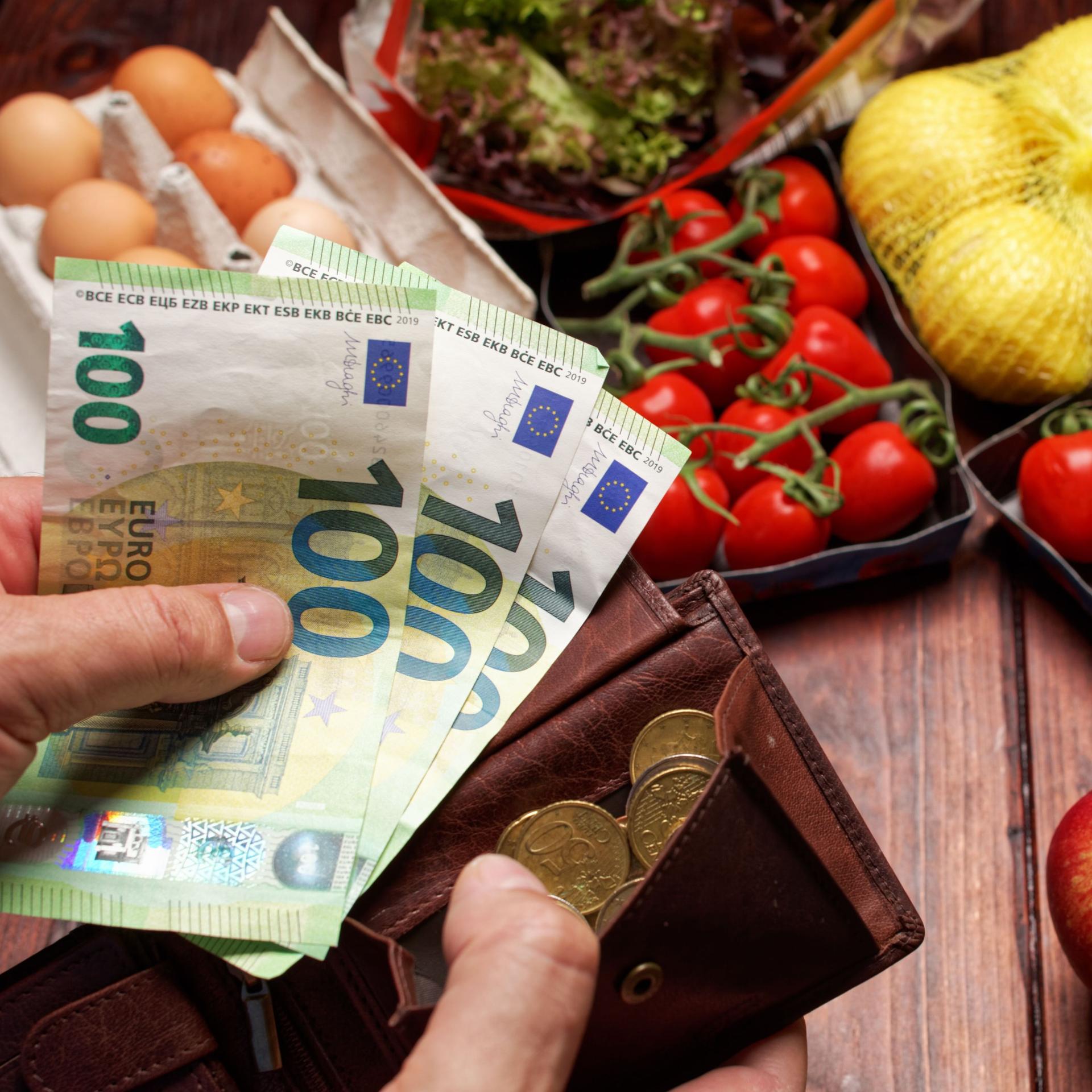 EIne Hand hält dre einhundert Euro-Scheine die andere Hand hält einen Geldbeutel im Hintergund liegt ein Einkauf mit Obst Gemüse und Eiern.